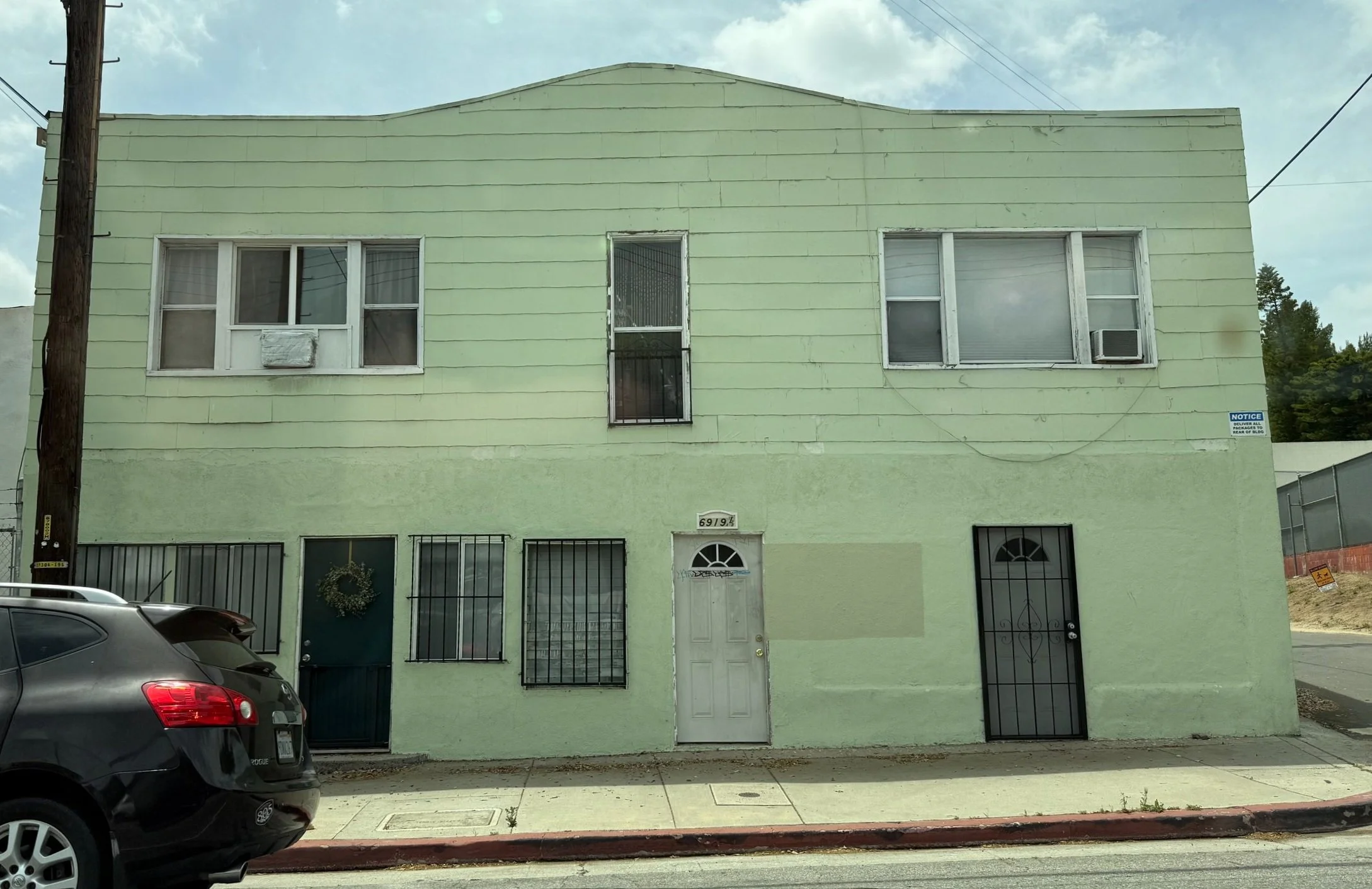 A two-story pale green building with barred windows and a white door with a semicircular window, located on a sidewalk with a parked black car in front. Windows on the upper level include an air conditioning unit and a screened window. There is a wooden utility pole on the left.