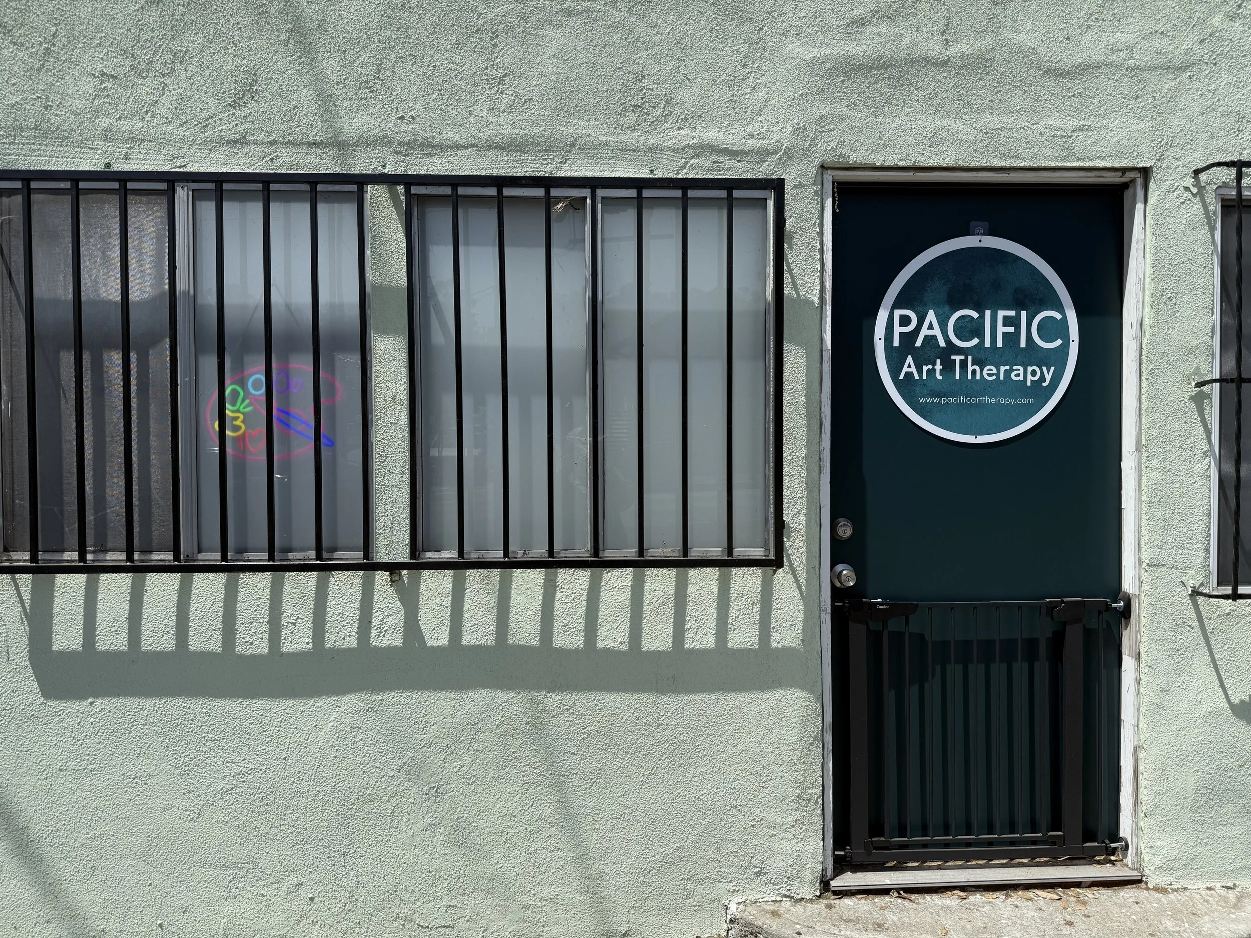 Building exterior with a window secured by black bars and a door labeled 'PACIFIC Art Therapy', featuring a circular sign with the website 'www.pacificarttherapy.com'; a small neon rainbow-colored art sign is also displayed in the window.