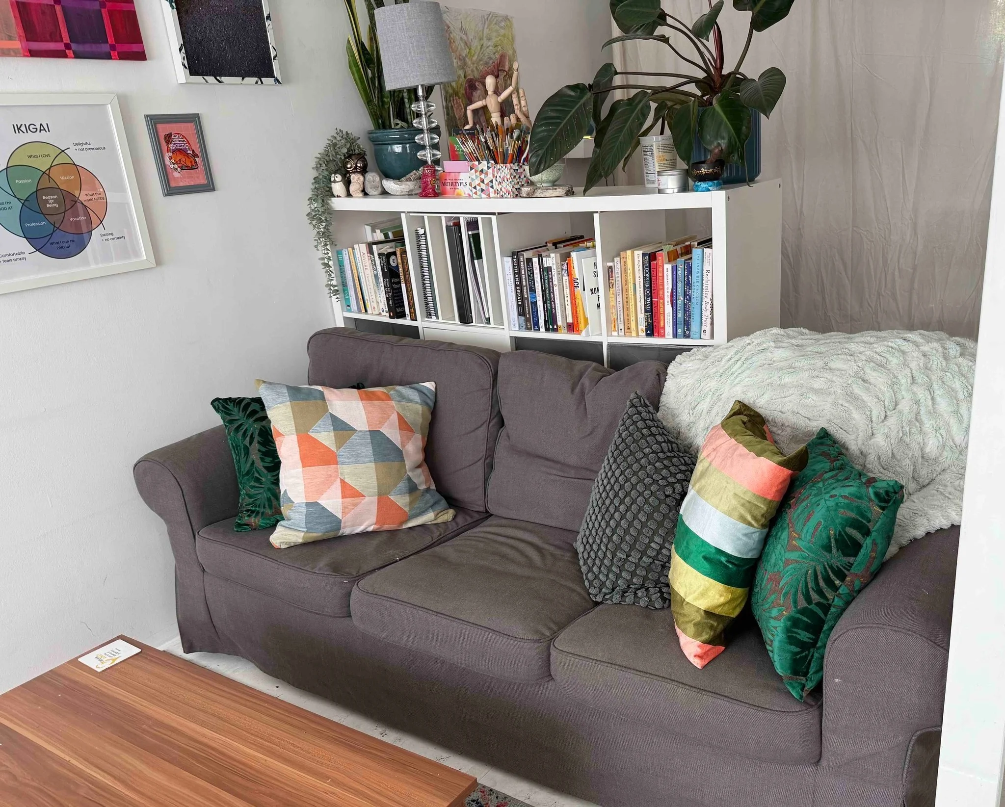 A living room with a gray sofa decorated with colorful patterned pillows. A white bookshelf filled with books and decorative items is behind the sofa. Green plants, a lamp, and a wall art with a circular diagram are on the wall. A wooden coffee table is in the foreground.
