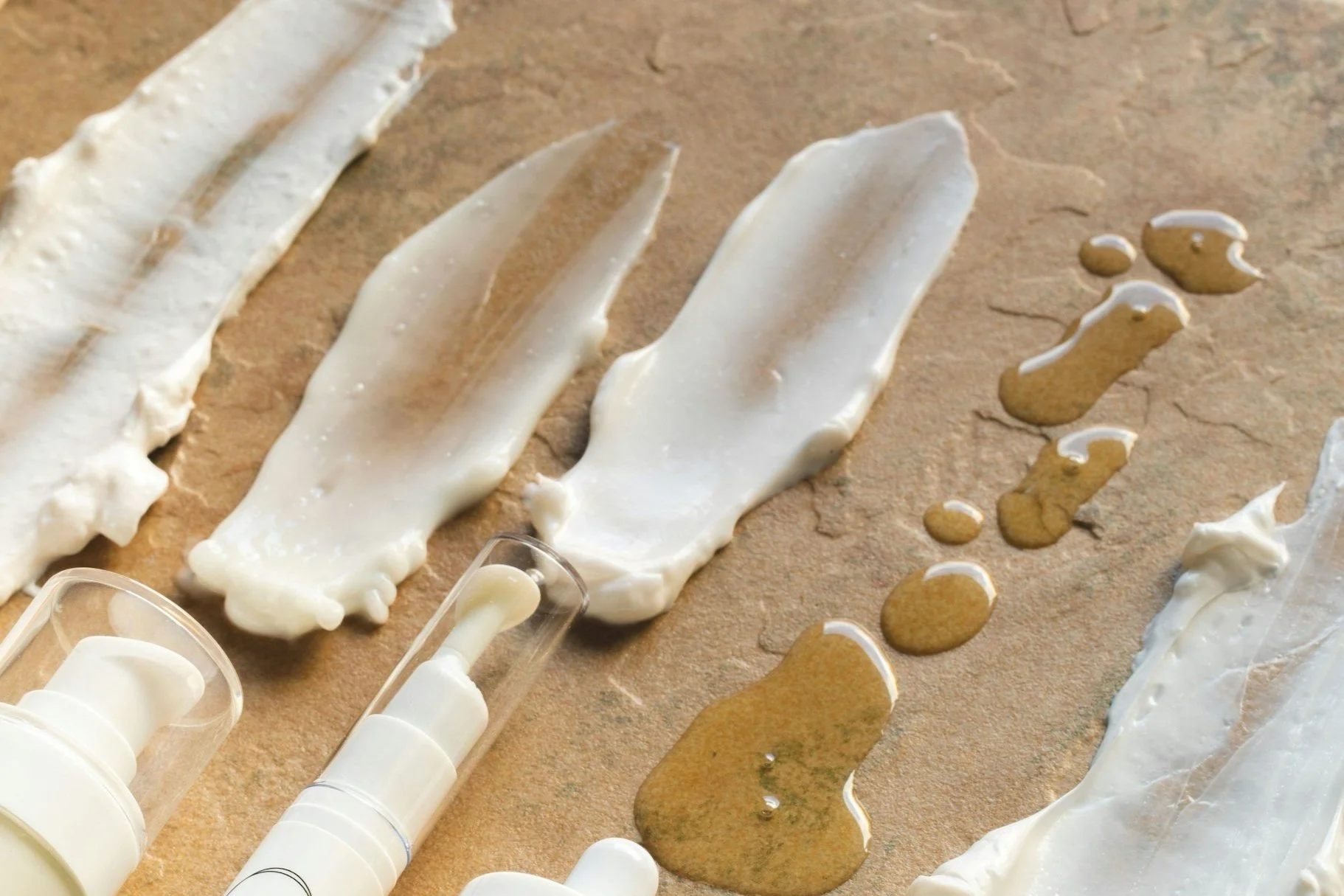 Various swatches of white cream or lotion and drops of golden liquid on a textured surface.