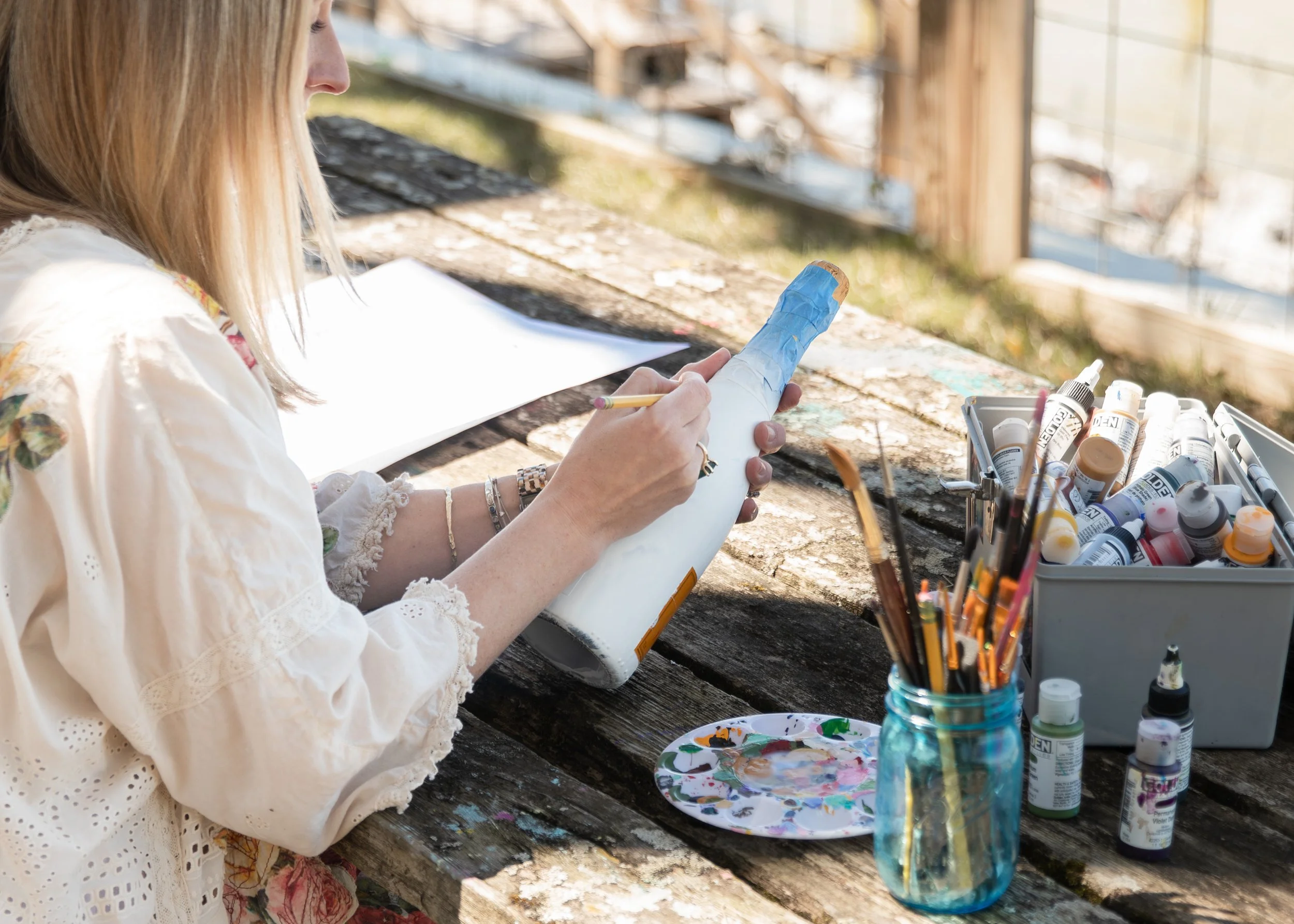 Marilu painting a glass bottle outdoors at a rustic wooden table with art supplies.