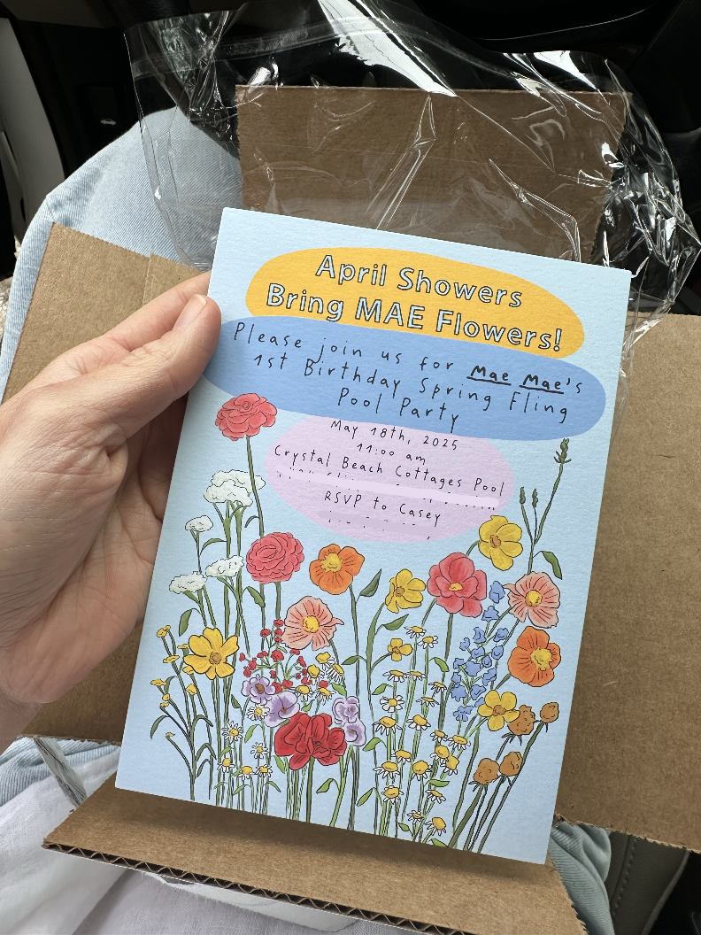 A person holding a colorful invitation card with floral designs, including poppies, daisies, and other wildflowers, for an April showers birthday pool party, scheduled on May 18th, 2025, at 11:00 am at Crystal Beach Cottages Pool, RSVP to Casey, with a plastic-wrapped gift box in the background.