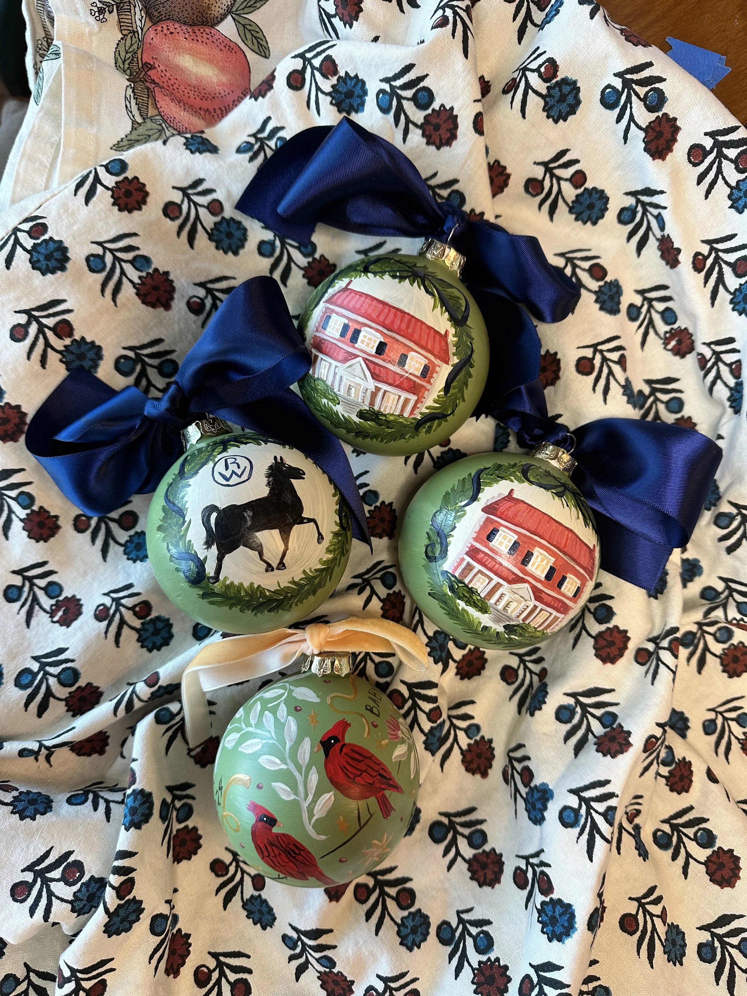 Four decorated Christmas ornaments with ribbons, placed on a fabric with a floral pattern. Two ornaments have house illustrations, one features a horse, and one shows red cardinals with greenery.