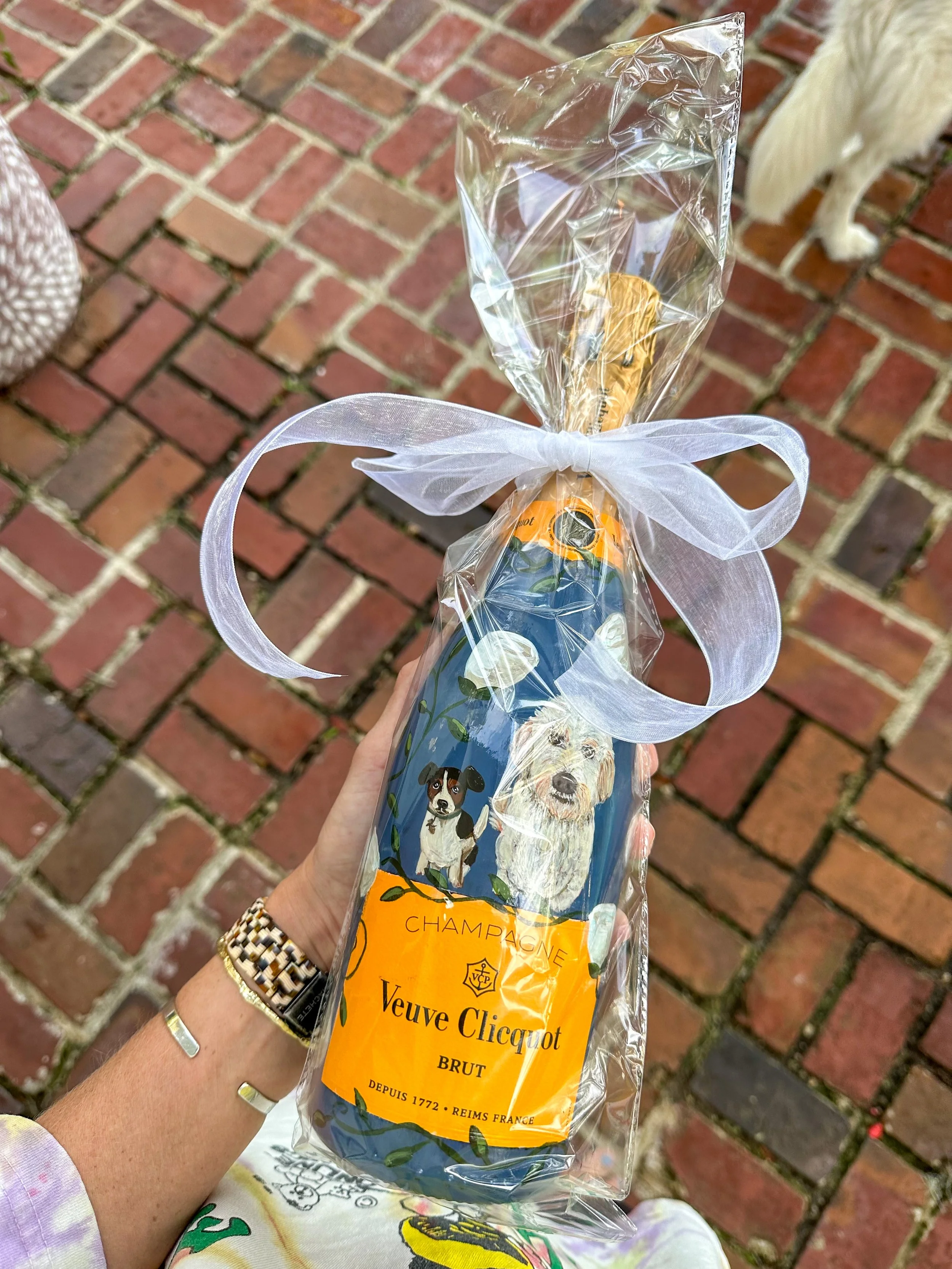 A hand holding a wrapped bottle of Veuve Clicquot champagne with cartoon dog illustrations, wrapped in clear plastic with a white ribbon, on a brick patio.