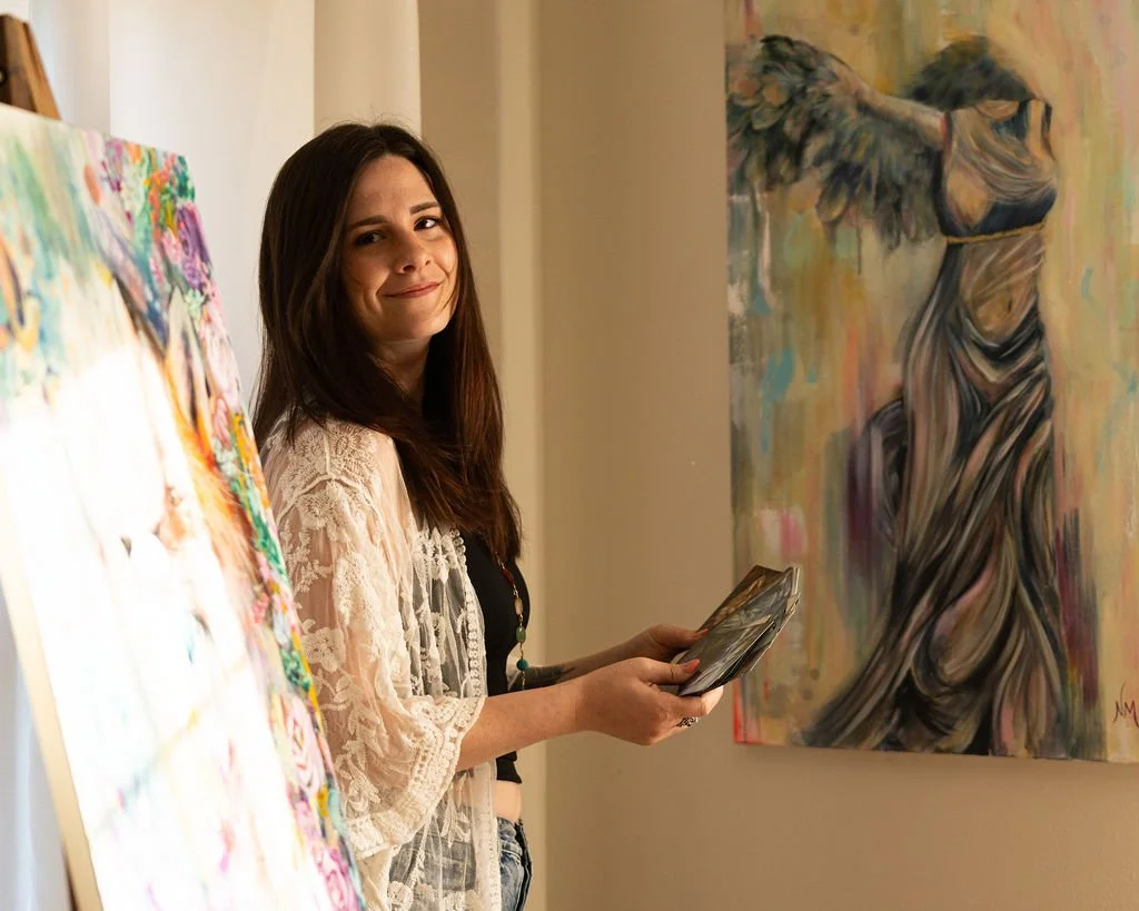 A young woman with long brown hair smiling at the camera while holding a tablet, standing next to colorful abstract paintings in an art gallery.