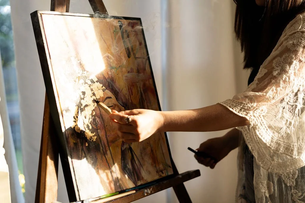 Woman painting abstract artwork on canvas, using a paintbrush, while holding a smartphone in her other hand.