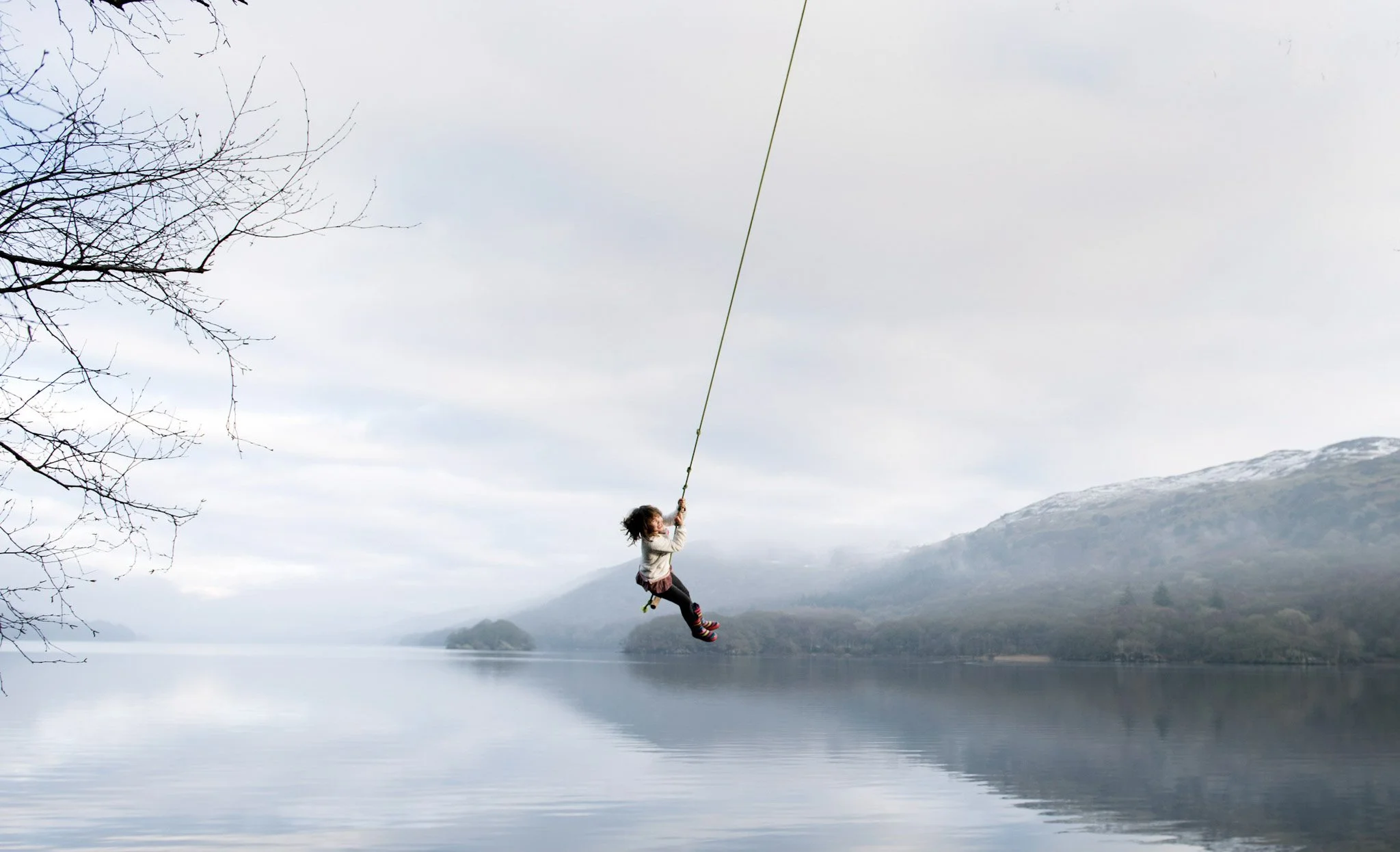 Child swinging on a rope over a calm lake with mountains in the background and a cloudy sky.