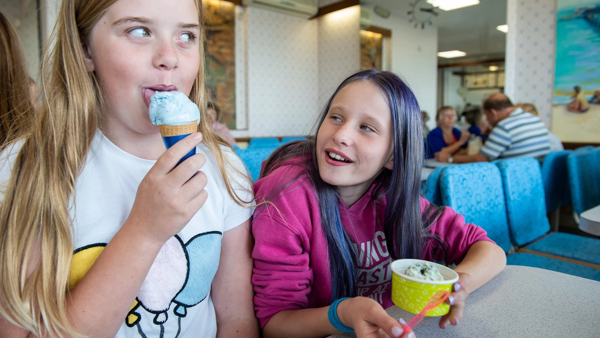 Two young girls enjoying ice cream at an ice cream shop, with other patrons dining in the background. Photography by Hannah Maule-ffinch.