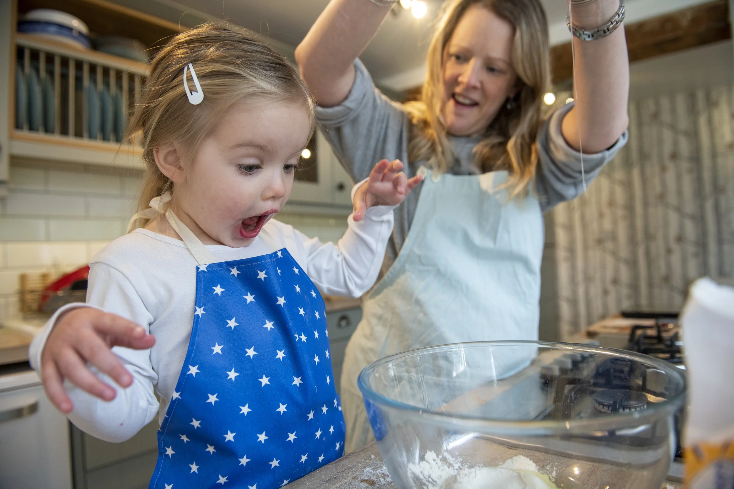 A young girl and an adult woman baking in a kitchen, with the girl expressing surprise as ingredients or dough are prepared on the countertop. Photography by Hannah Maule-ffinch.