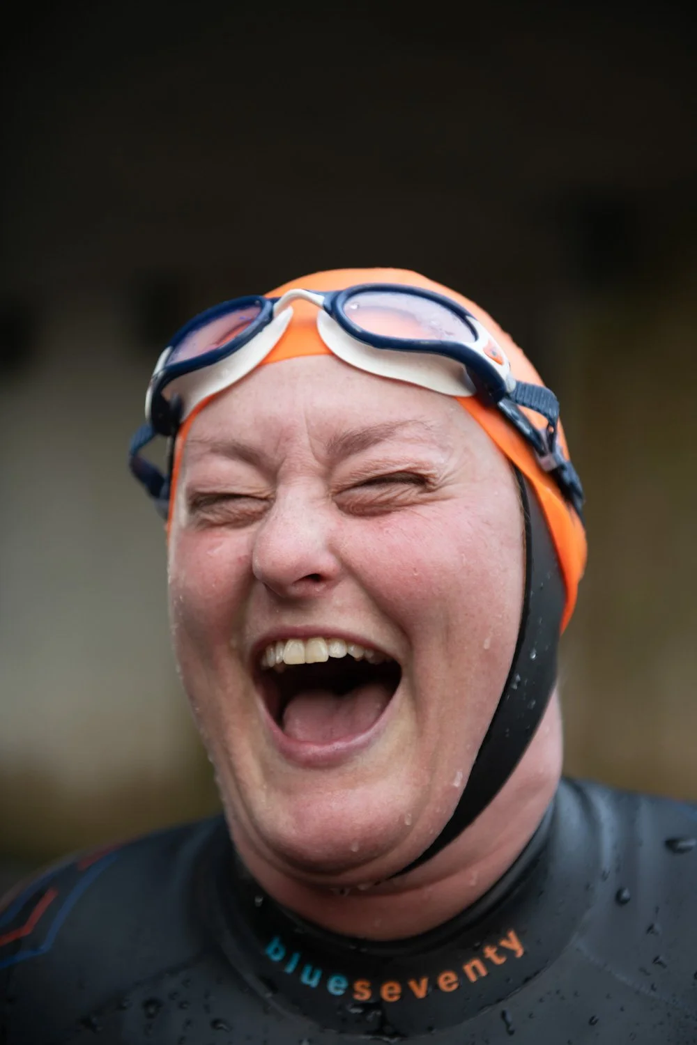 Close-up of a woman in swimming gear, wearing an orange swim cap and goggles on her forehead, cheering or shouting with her eyes closed. Photography by Hannah Maule-ffinch.