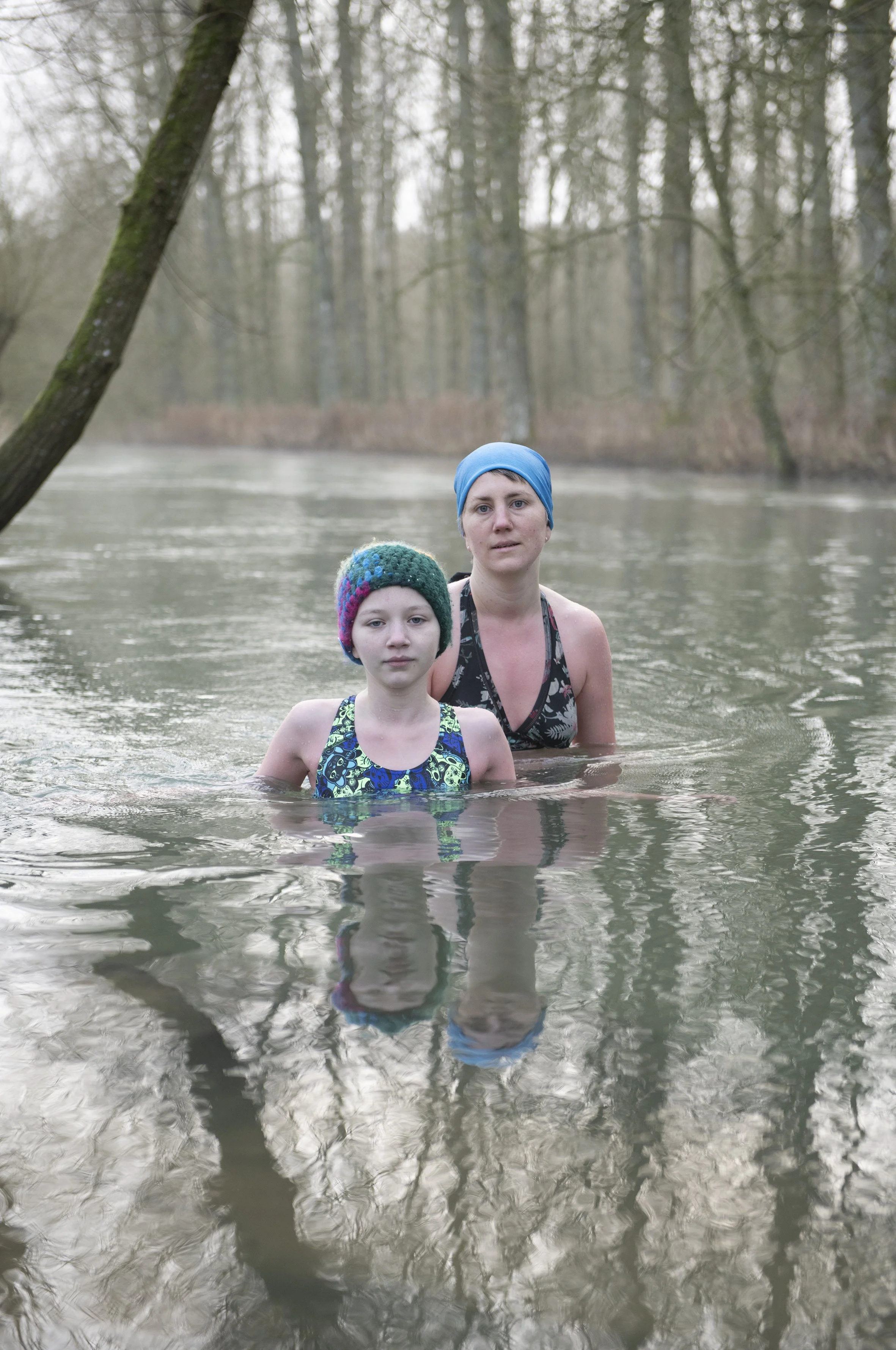 Two women, one young girl and one adult, standing in a river with trees in the background, wearing swim caps and swimsuits.