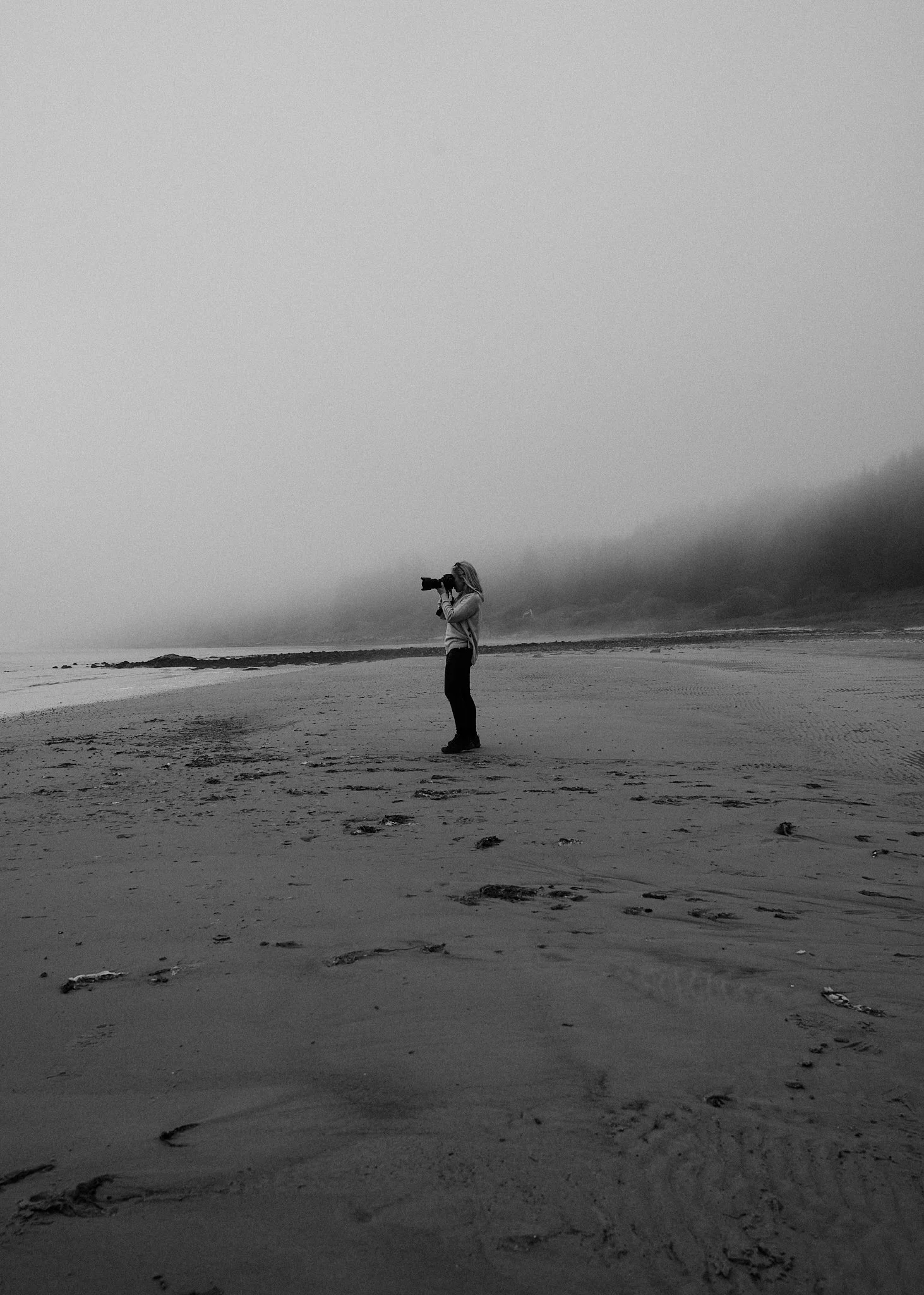 Hannah standing on a beach, holding a camera to their eye, taking a picture, with fog in the background.
