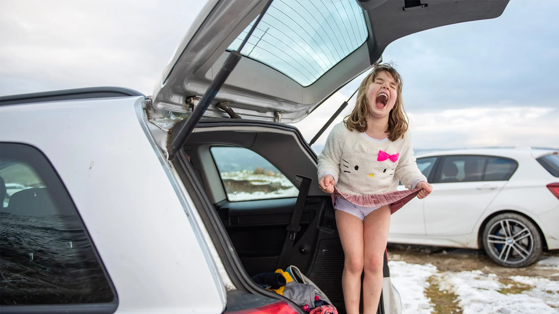 A young girl with a Hello Kitty sweater and pink bow on it, standing in the trunk of a white car, laughing and holding her skirt in a playful manner, with cars and snow on the ground in the background.