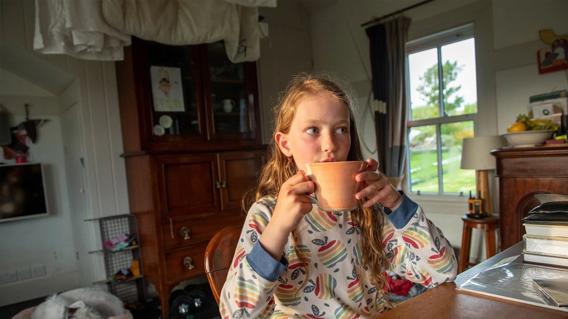 A young girl with long curly hair sipping from a ceramic cup at a wooden table in a cozy room with a large window showing a green outdoor landscape.