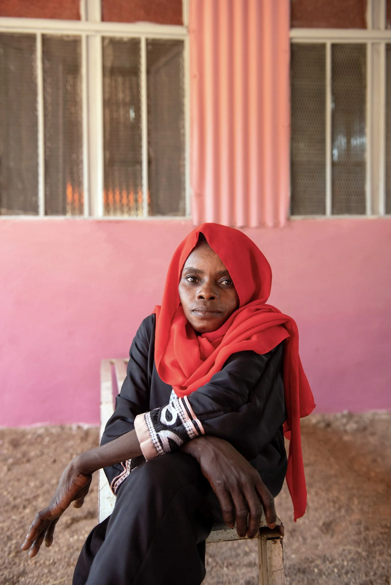 A woman sitting on a chair in front of a pink wall with large windows, wearing a black outfit and a red headscarf.