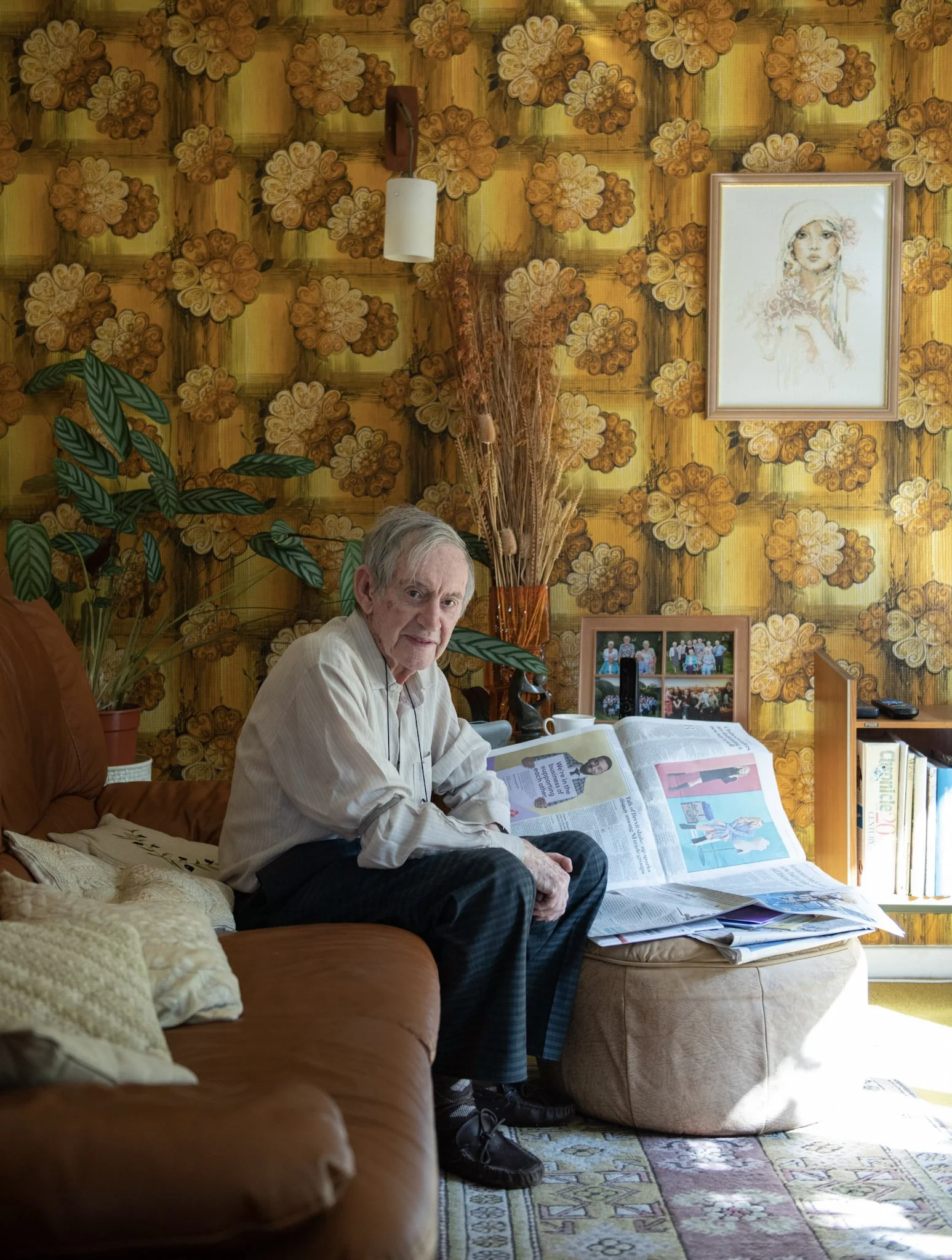 An elderly man in a white shirt and plaid pants sitting on a brown couch in a cozy living room, reading a newspaper placed on an ottoman. The room features a vibrant yellow wallpaper with orange floral patterns, a framed sketch of a woman on the wall