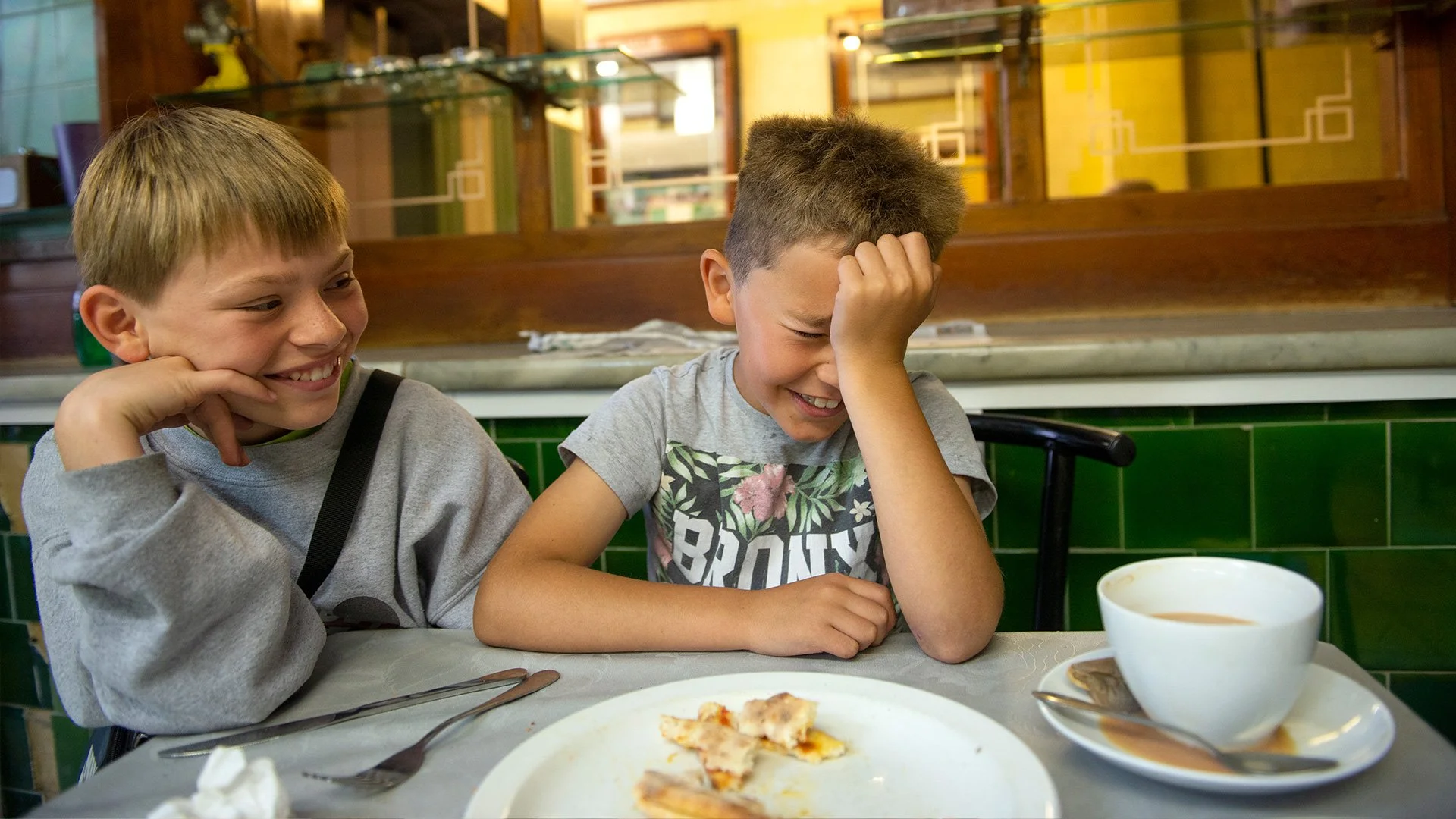 Two boys sitting at a table in a restaurant, laughing with one boy holding his head in his hand and the other smiling. There is a partially eaten pizza on a plate, a cup of coffee, and a spoon on the table.
