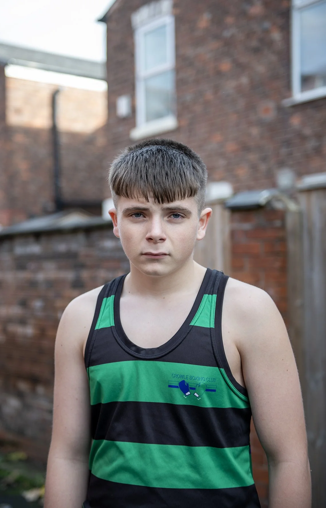 A young man with short brown hair and blue eyes stands outdoors in front of a brick building with windows. He is wearing a black and green striped sleeveless athletic shirt with a boxing club logo. Photography by Hannah Maule-ffinch.
