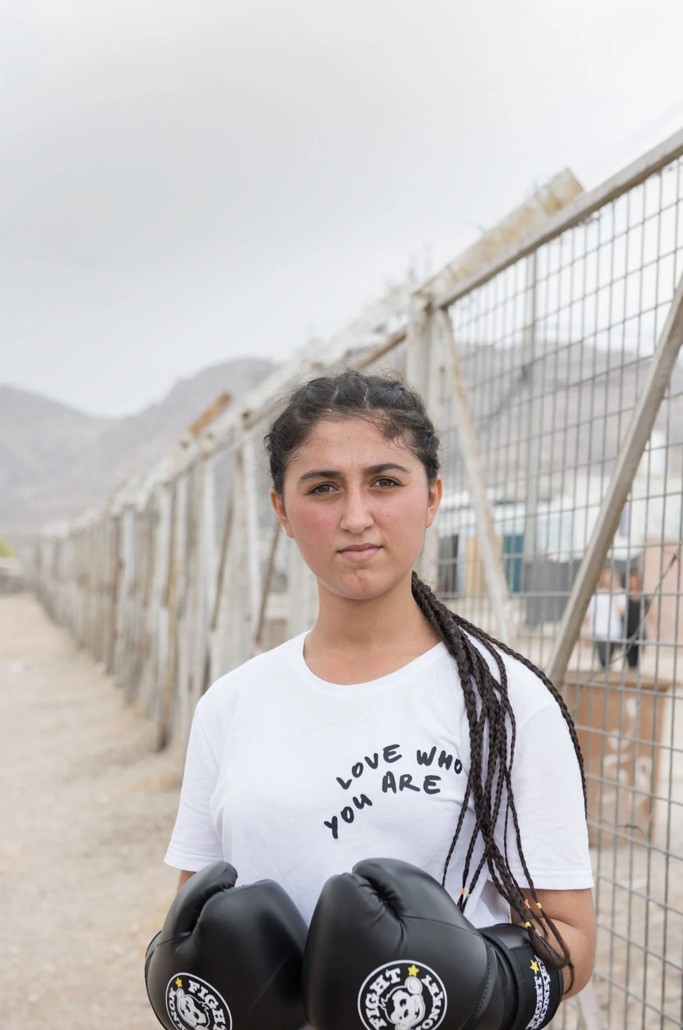 A young woman with boxer gloves standing outdoors next to a fence, wearing a white t-shirt that says 'Love Who You Are', with mountains in the background.