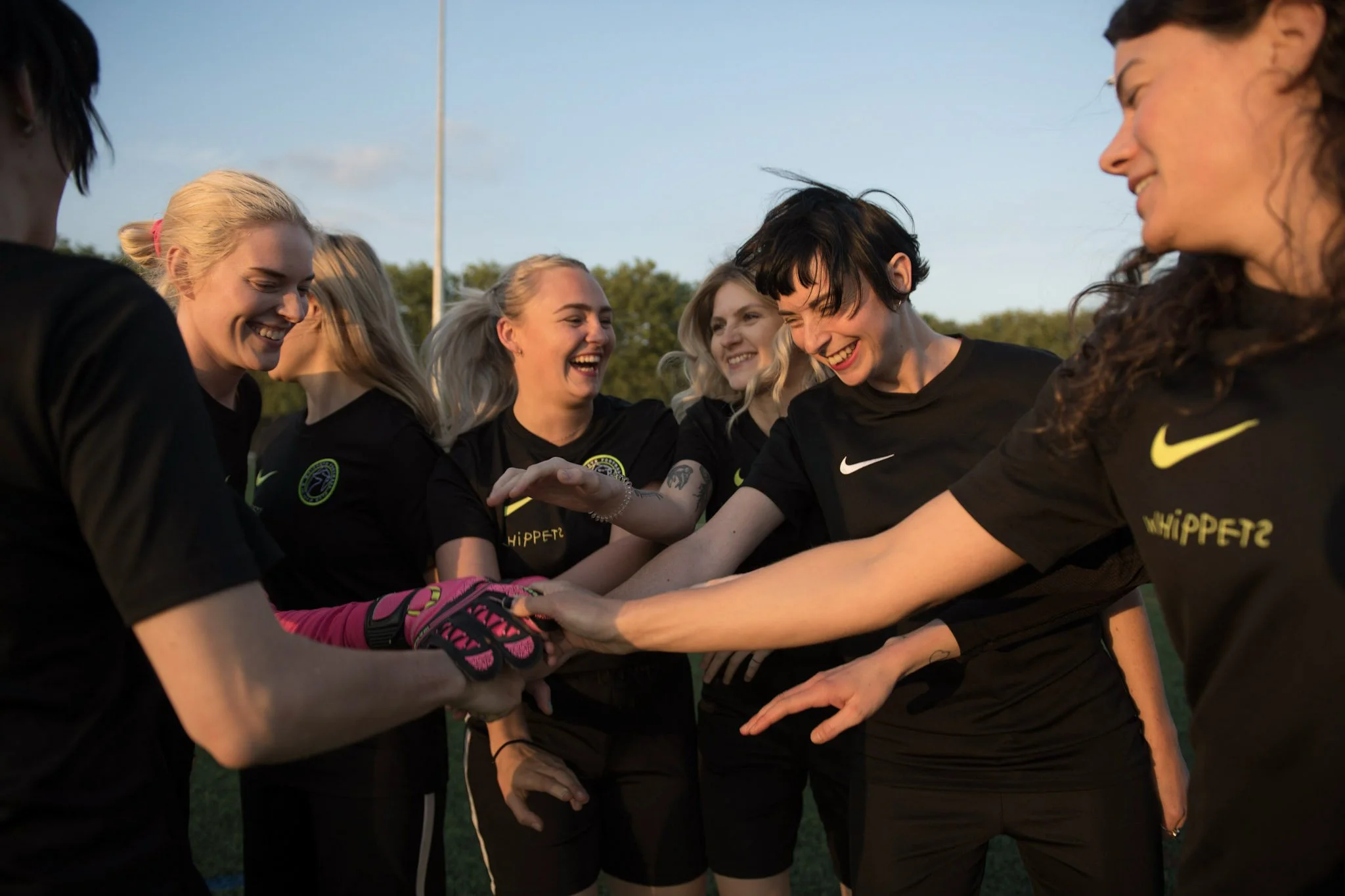 Group of female soccer players wearing black Nike jerseys with yellow accents, smiling, and high-fiving outdoors during the daytime.