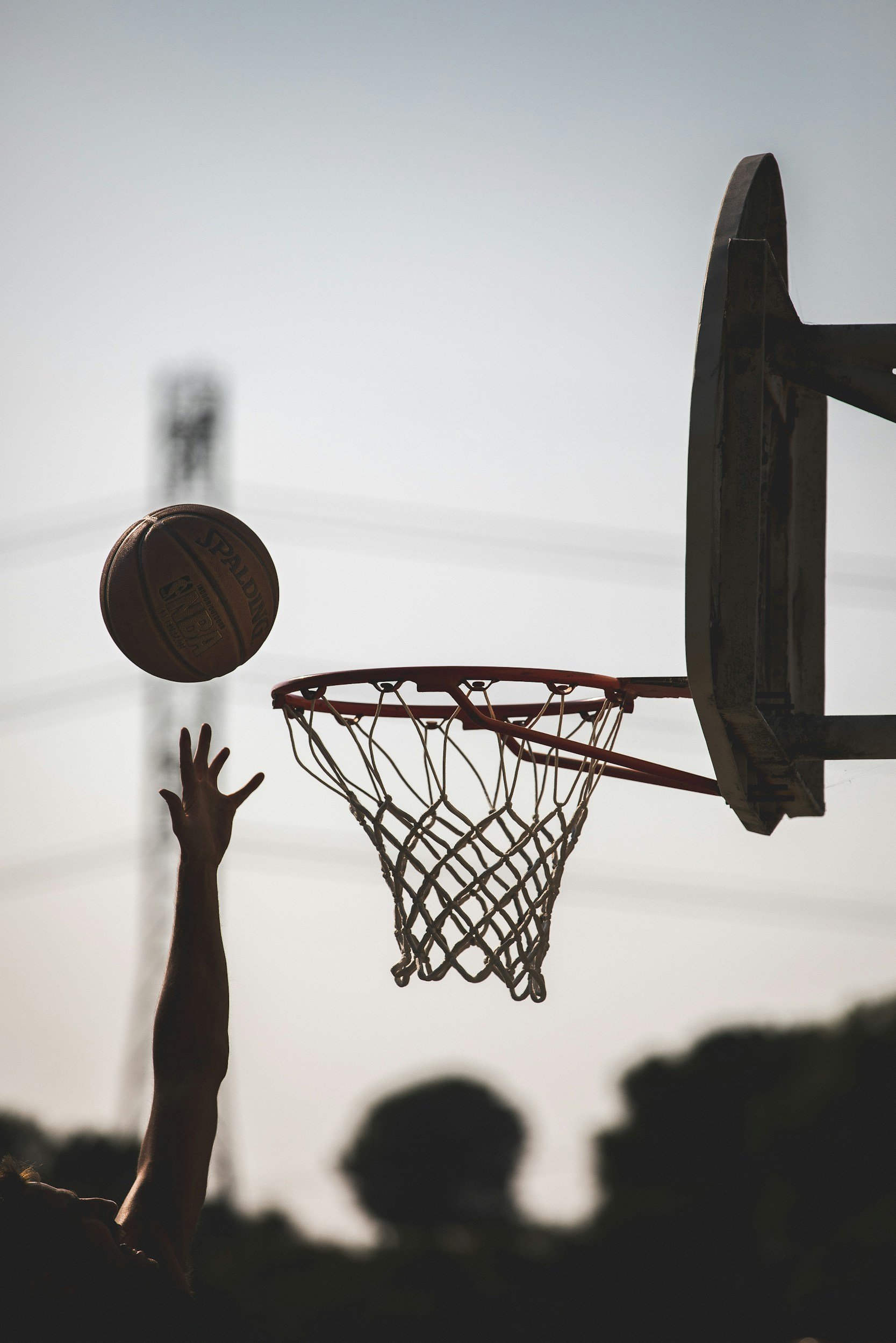 A person reaching up with an arm extended towards a basketball near a hoop, with the basketball in midair close to the hoop, during sunset or dusk outside.