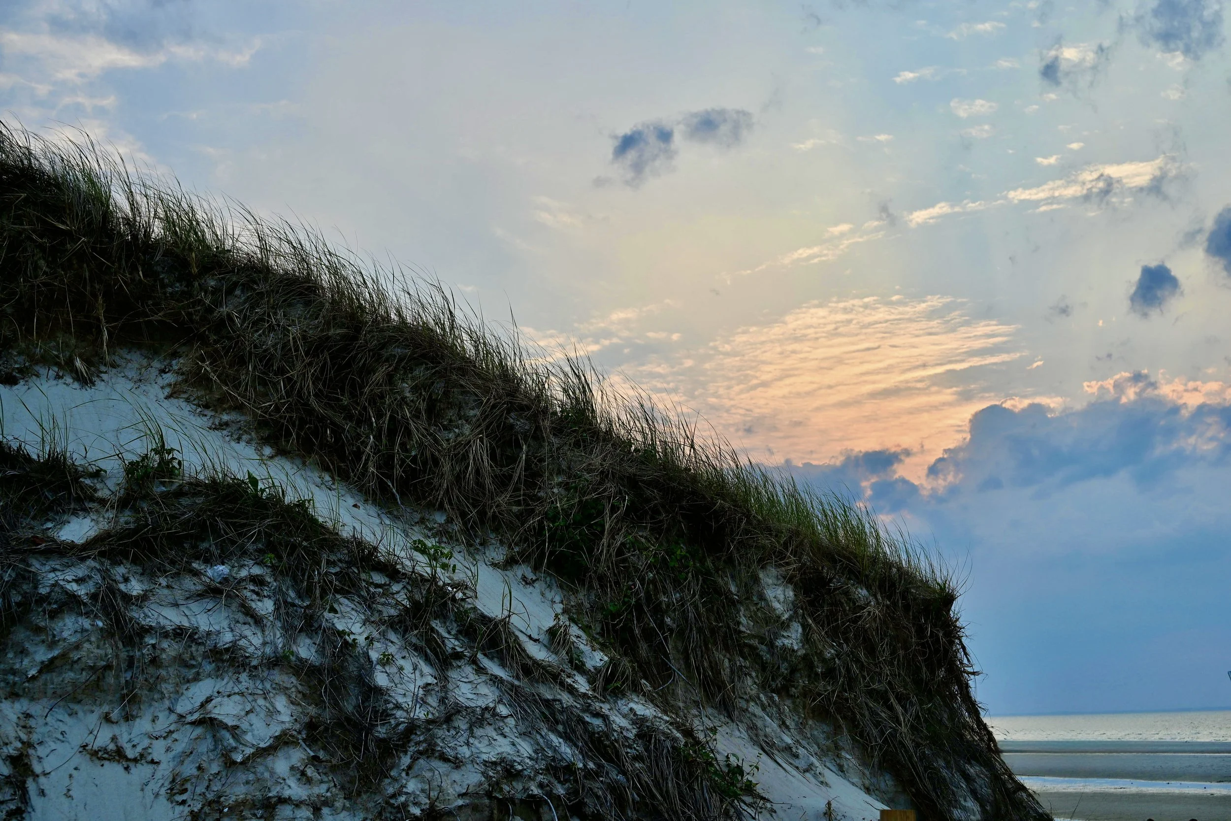 A sandy dune covered with grass next to a beach with the ocean in the background, under a partly cloudy sky at sunset.