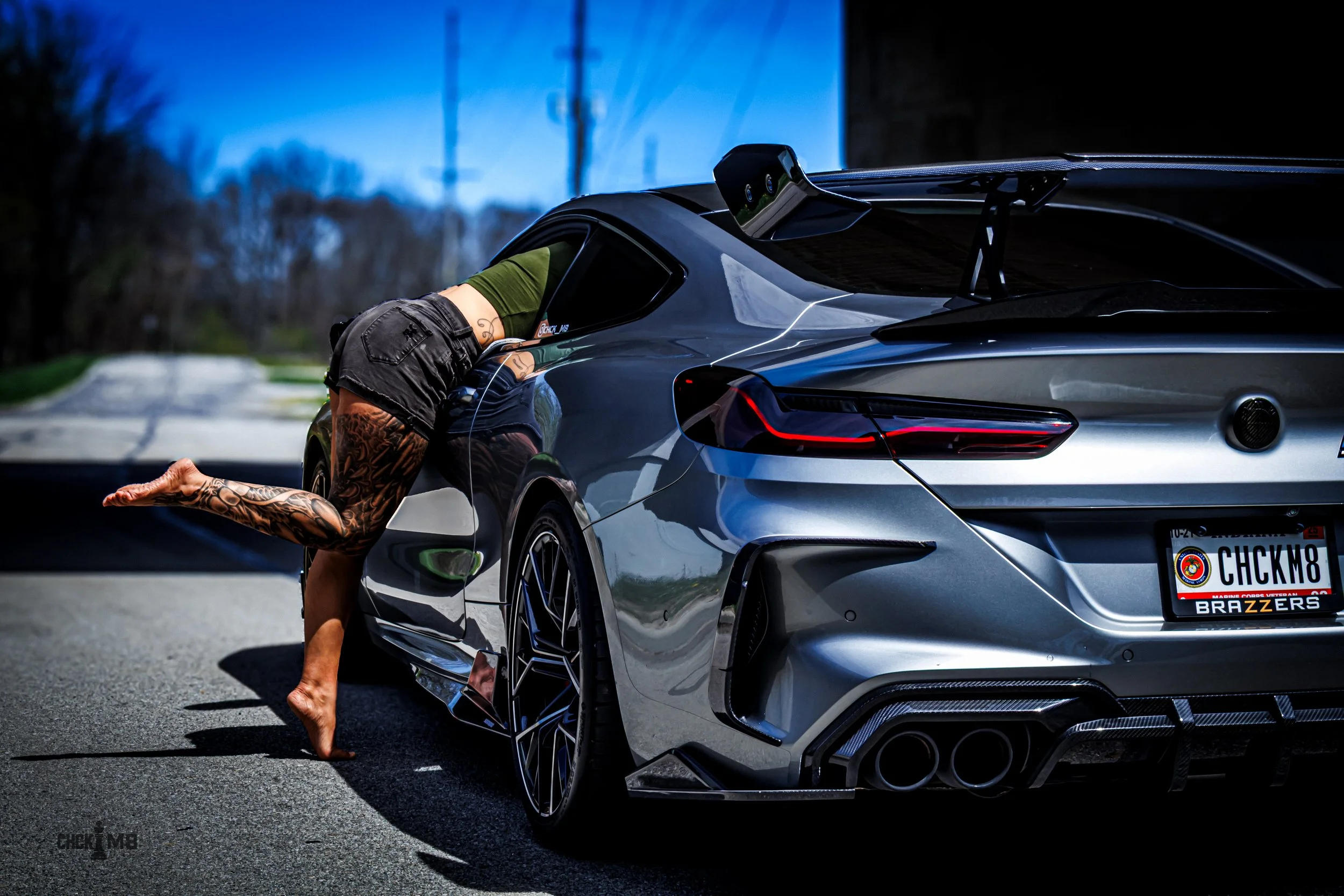 A person with tattoos wearing shorts and a green shirt lies in a bent-over pose with their head and upper body inside the open window of a silver luxury sports car on a suburban street. The car has a custom license plate reading 'CHCKM8' and visible branding for 'BRAZZERS'.