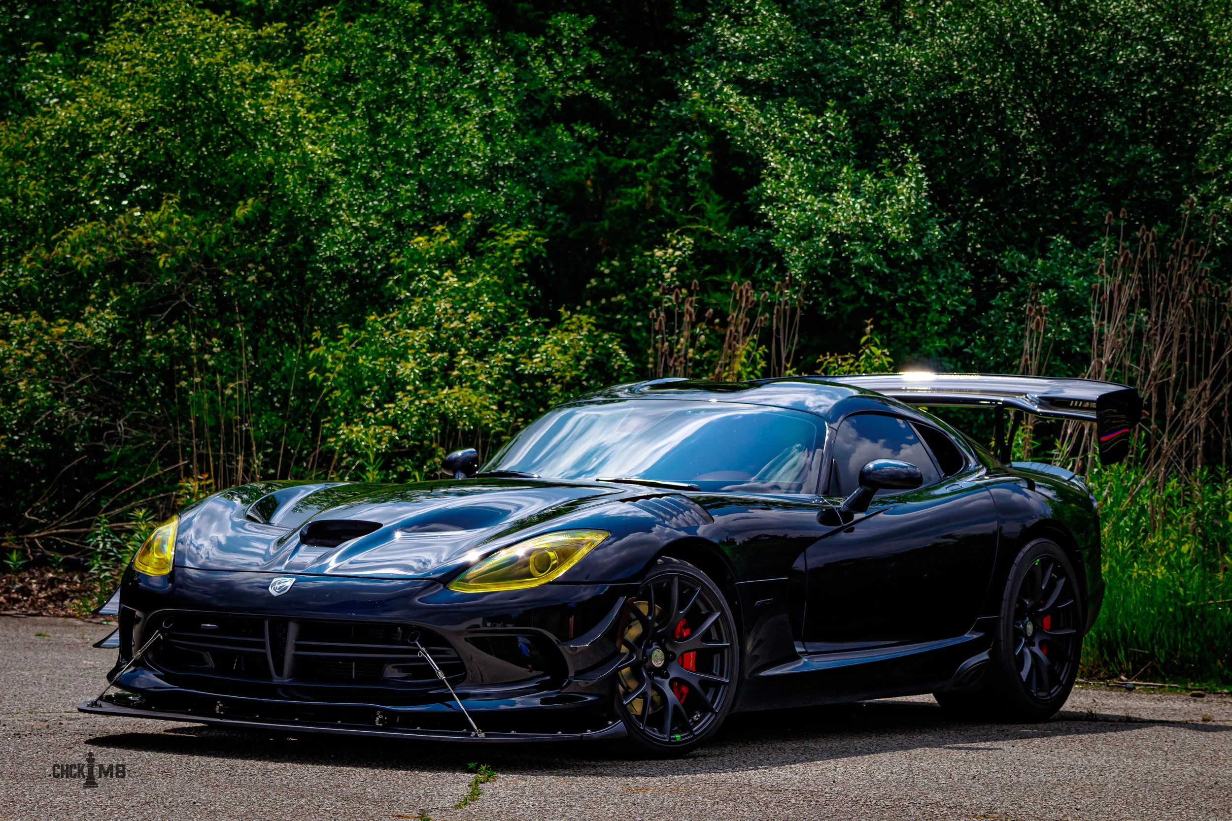 Black sports car with a large rear spoiler parked on a paved road, greenery in the background, yellow tint on headlights, and a front lip spoiler.