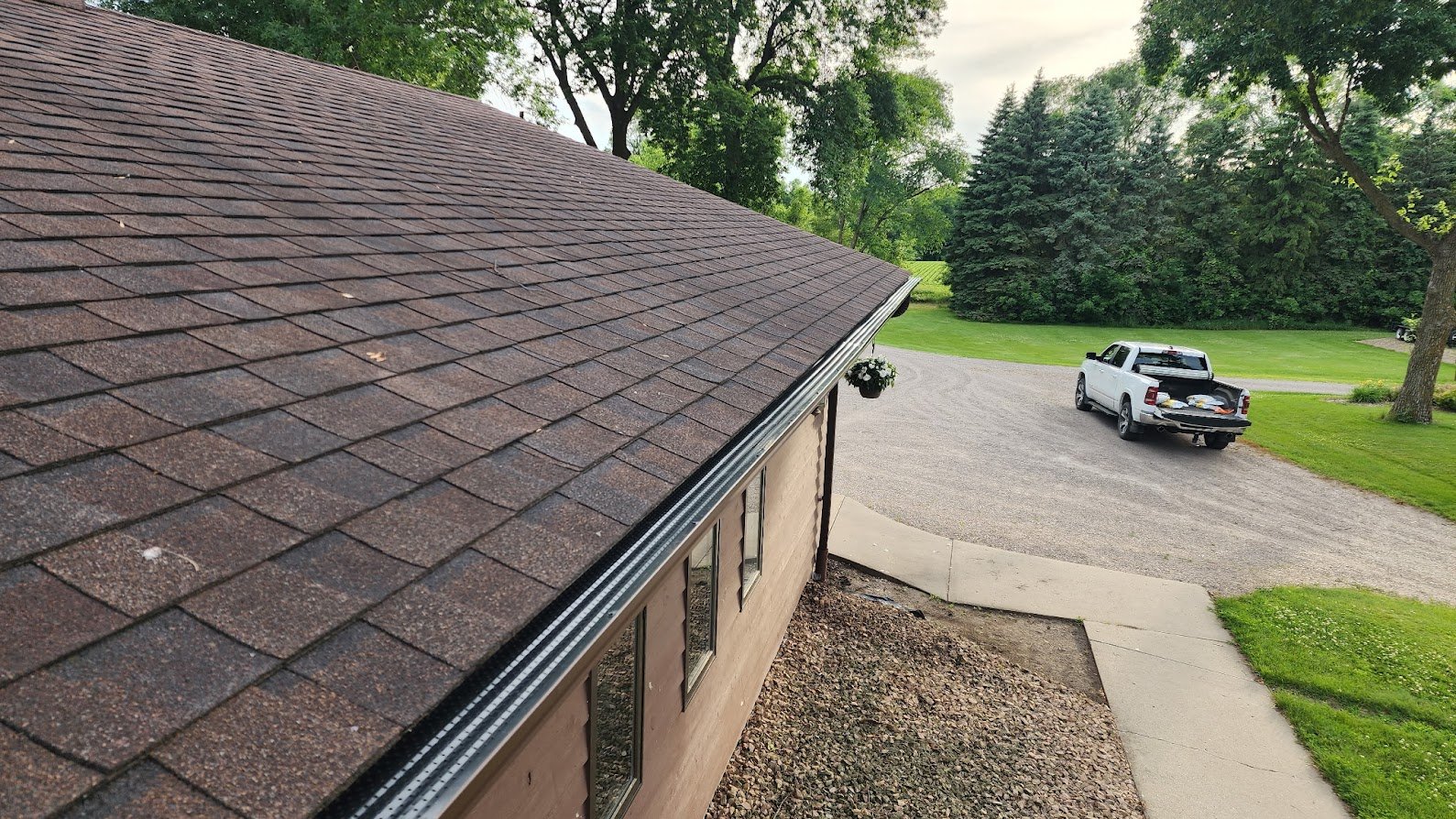 View from the side of a house showing a brown shingle roof, a window, and a hanging flower pot, with a driveway, a white pickup truck, and green trees and lawn in the background.