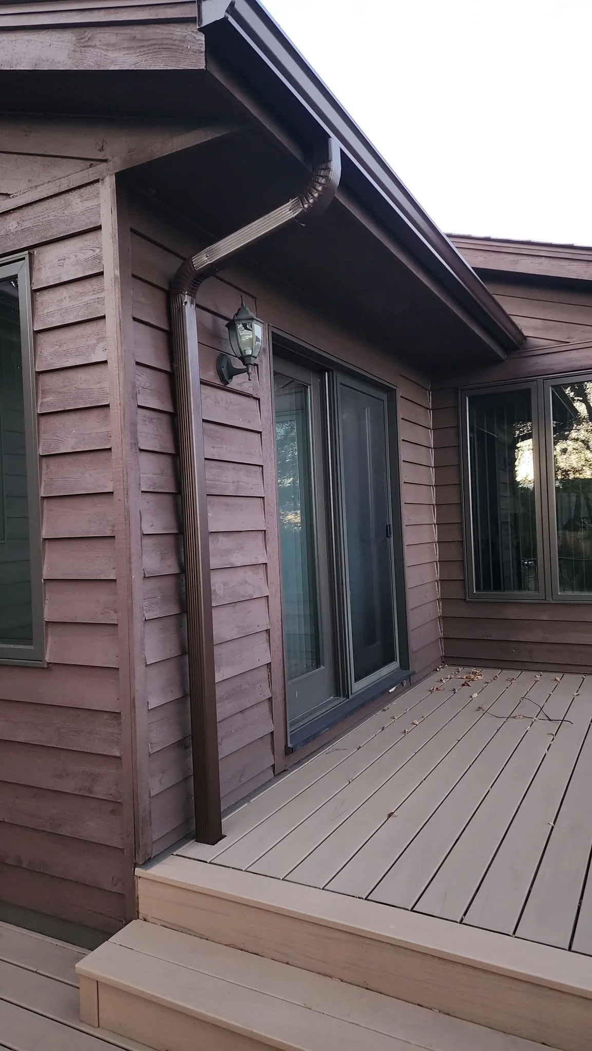 A wooden deck outside a house with sliding glass doors, a window, and an exterior wall with wood siding. There is a gutter and a wall-mounted outdoor light fixture.