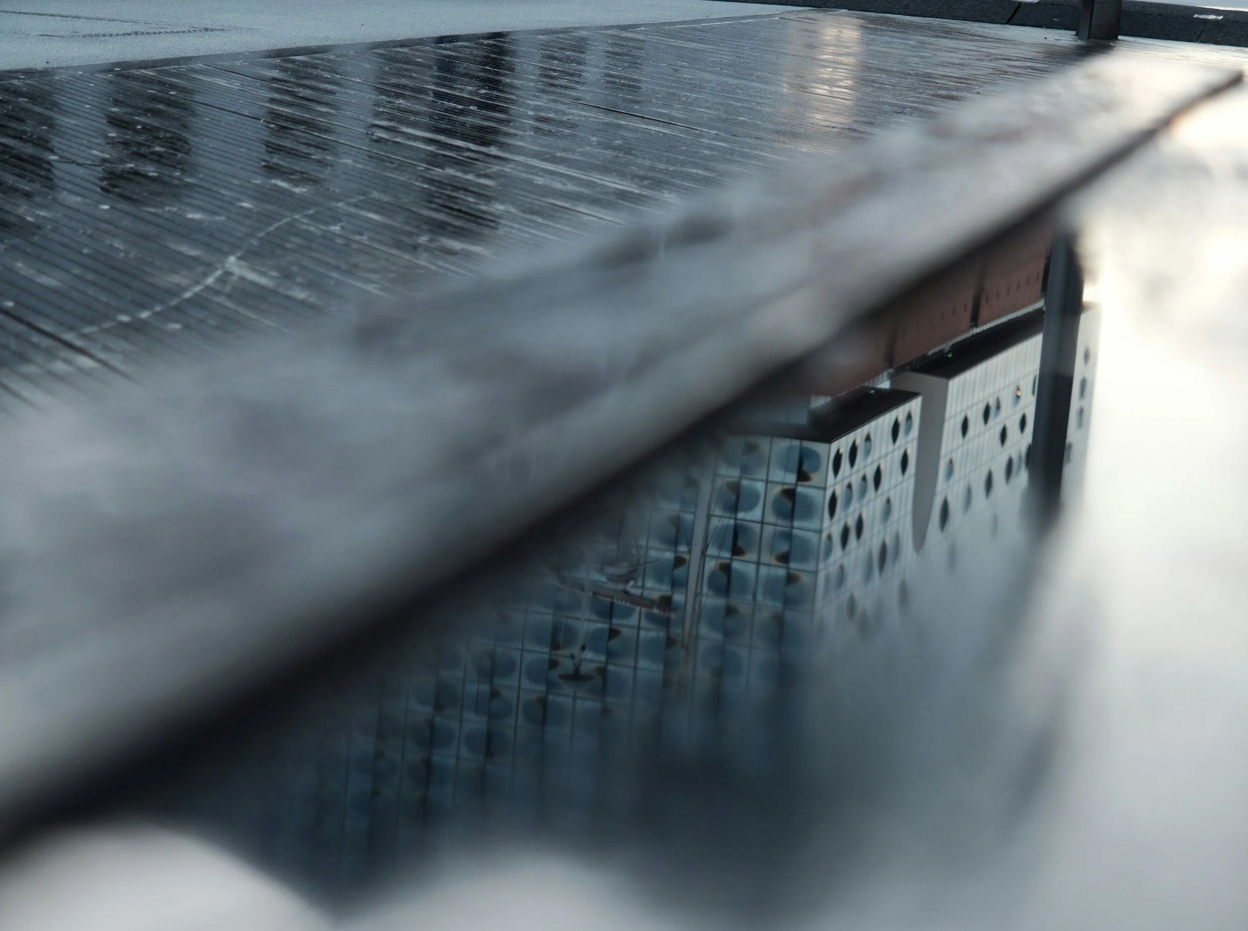 Close-up of a solar panel with reflections and raindrops on its surface, mounted on a structure with a building in the background.