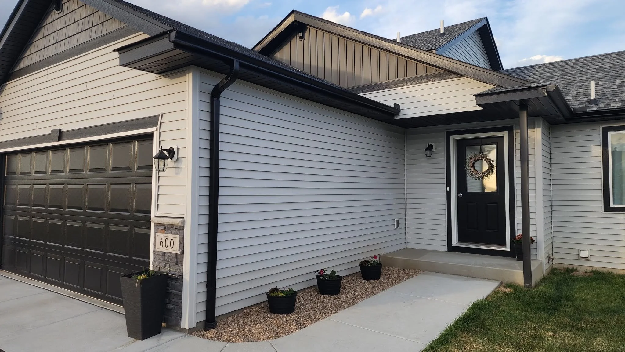 Front exterior of a modern house showing a black garage door, a white house with black accents, a front door decorated with a wreath, potted plants, and a driveway with grass and concrete sidewalk.