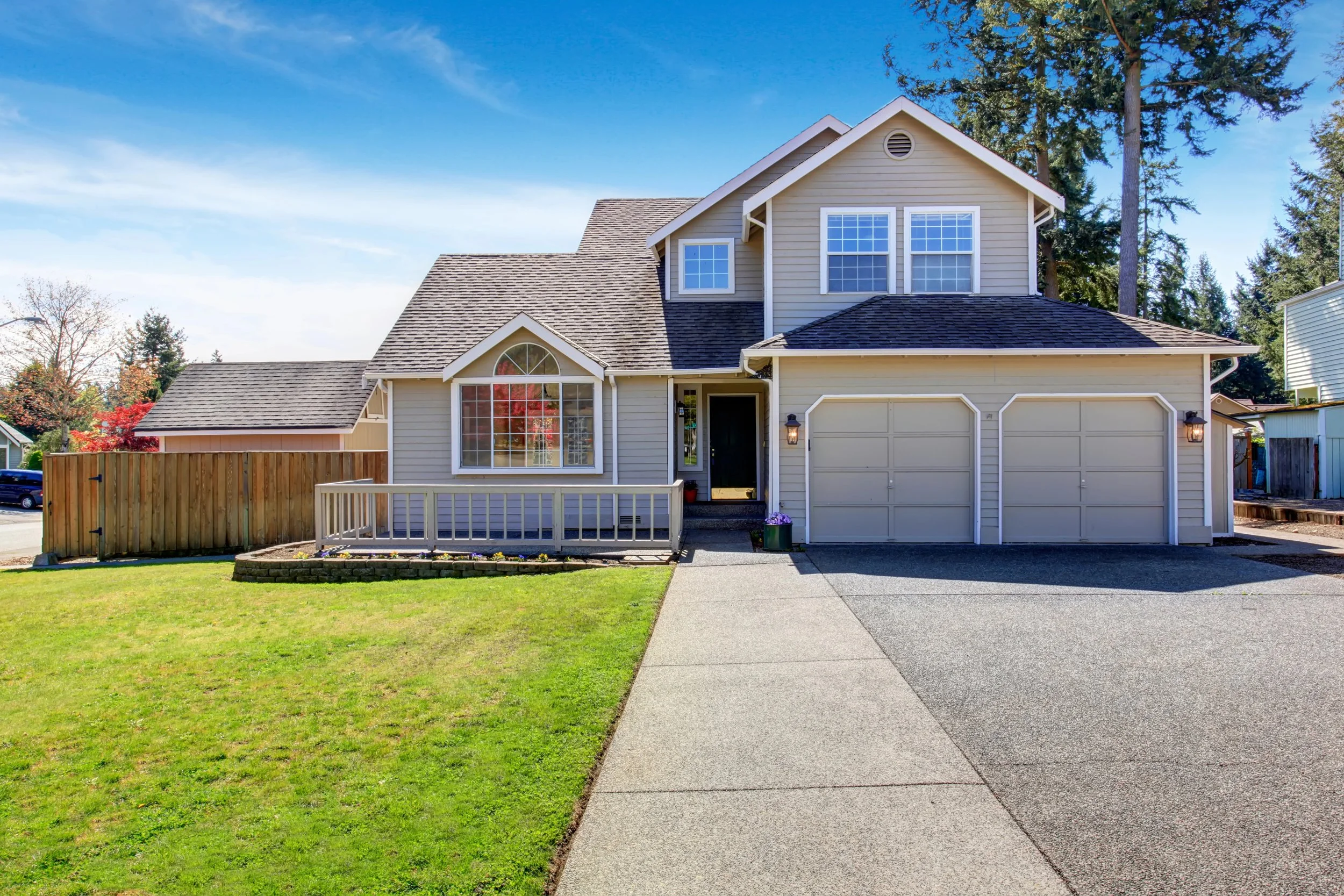 Front view of a two-story house with beige siding, large front window, attached garage, and a small front porch with a white railing. A concrete driveway leads up to the garage, and there is a grassy yard with a wooden fence on the left side. Tall trees are in the background under a bright blue sky with a few clouds.