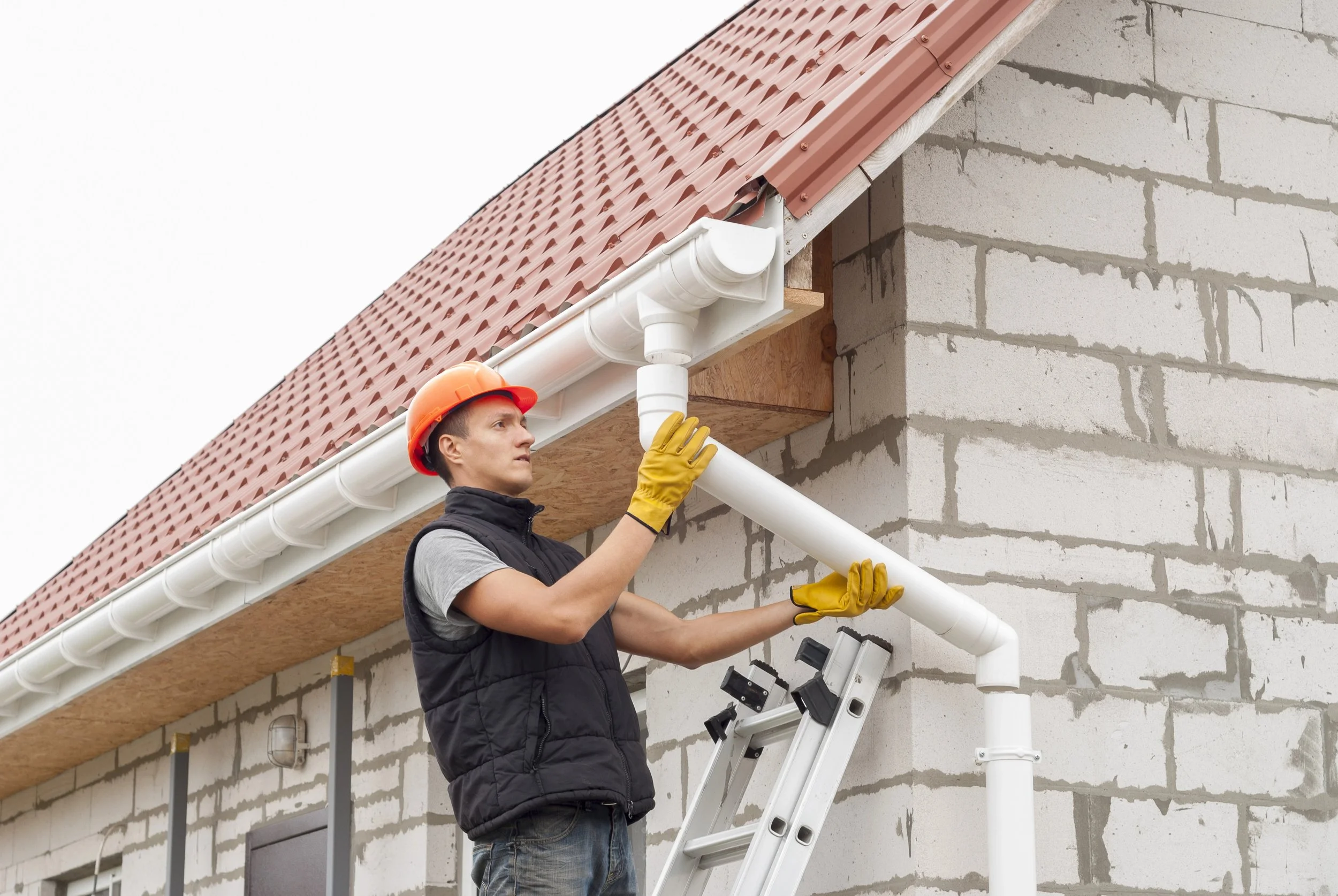 A man wearing a red hard hat and yellow gloves installing gutter pipes on the side of a brick house.