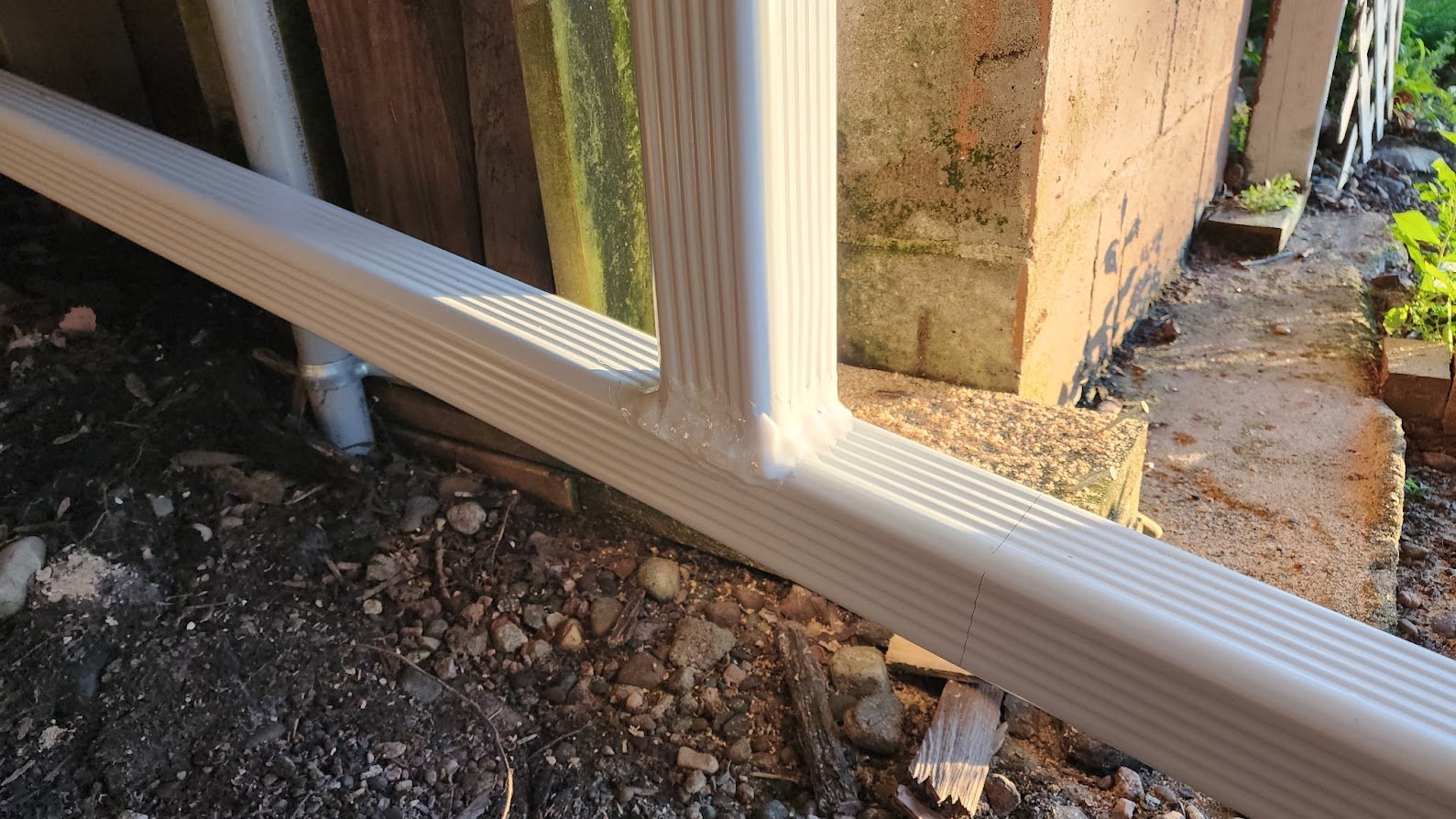Close-up of white plastic or vinyl drainage pipe and downspout at the corner of a house, with soil and rocks on the ground, and part of a brick wall and wooden fence visible.