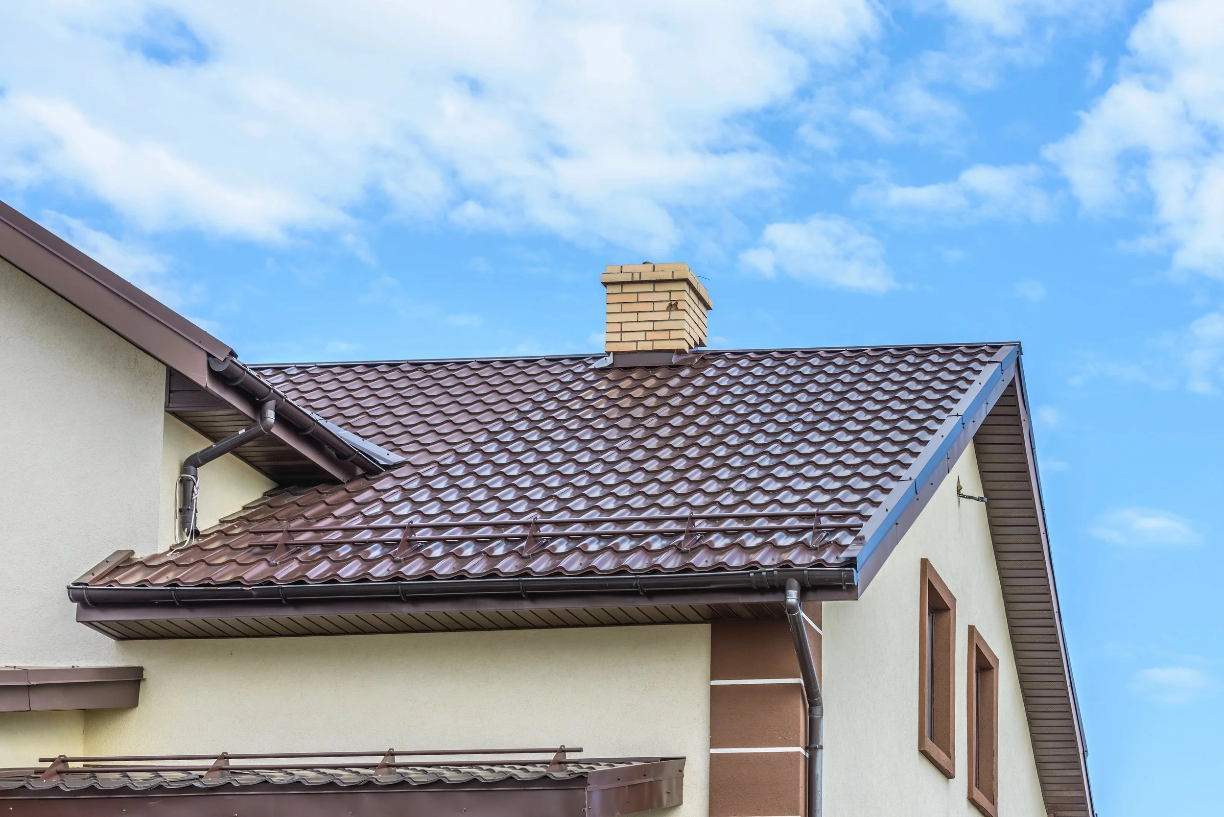 A house with a beige brick chimney, a brown tiled roof, and a rain gutter against a bright blue sky with some scattered white clouds.