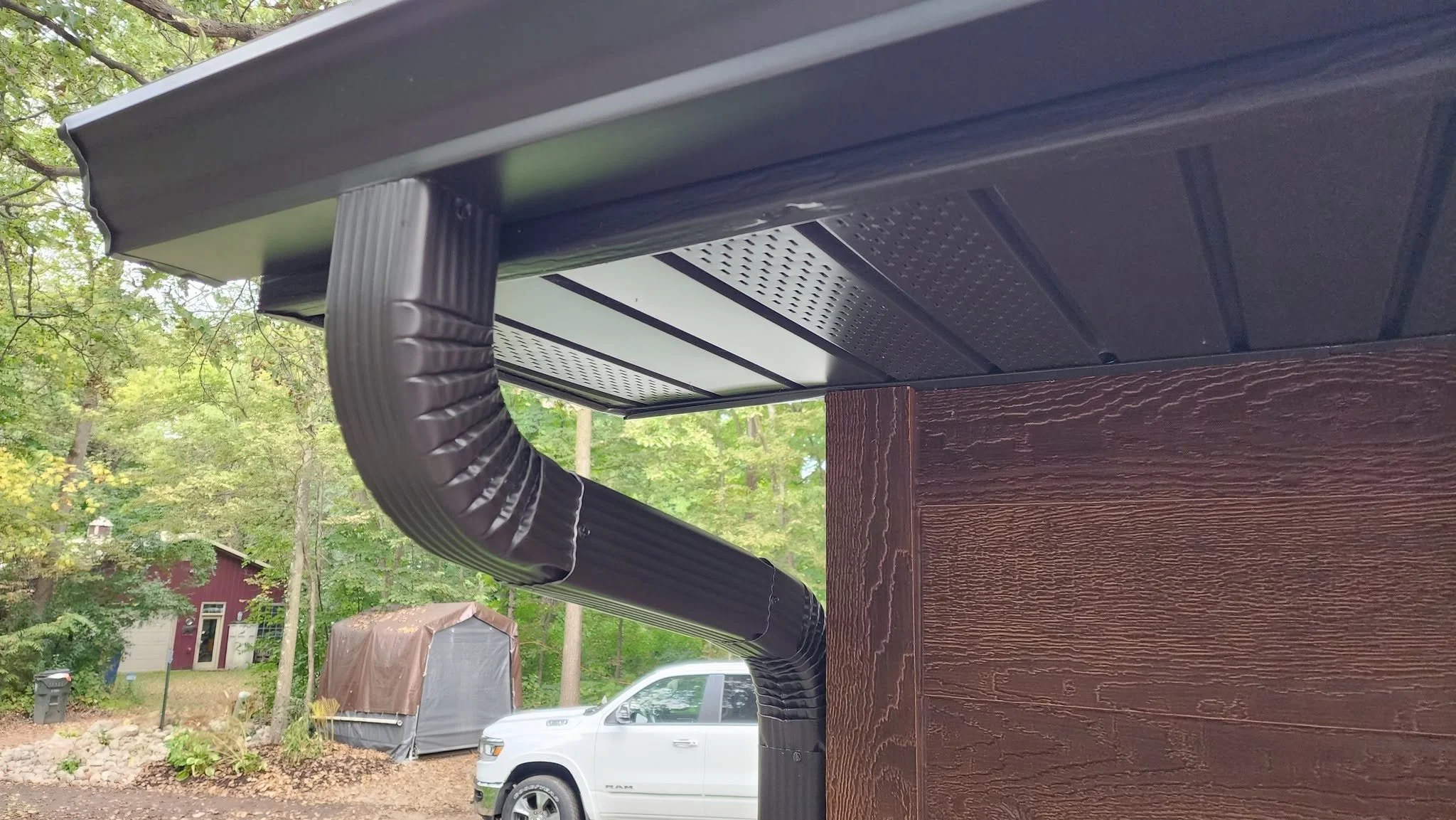 Close-up of a black downspout and gutter attached to a wooden structure, with a background of trees, a white vehicle, and a shed with a tarp covering on a yard.