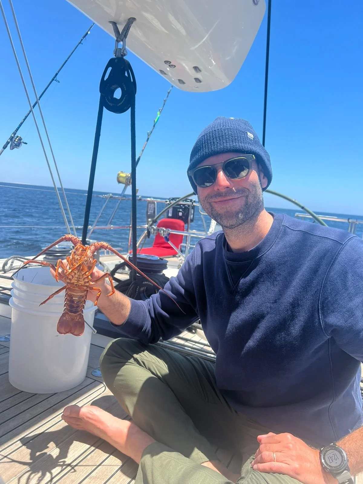 A man sitting barefoot on a boat deck, holding a lobster over a white bucket, with the ocean and clear blue sky in the background.