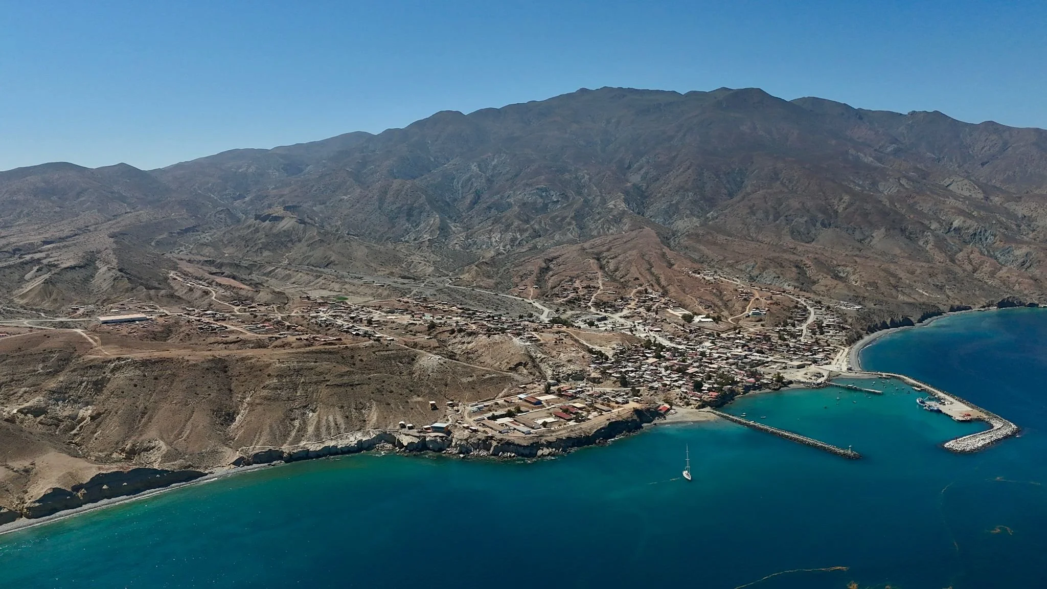 Aerial view of a coastal town with mountains in the background, a harbor with a breakwater, and turquoise water.