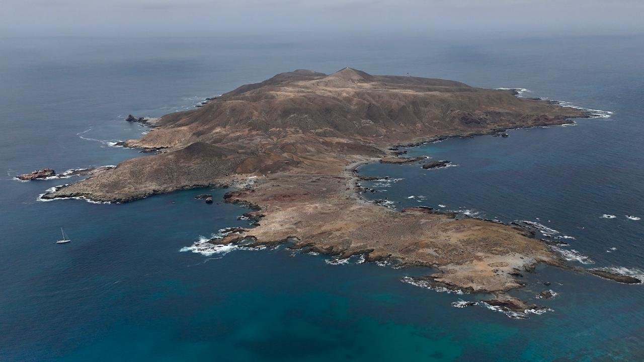 Aerial view of a rocky, barren island surrounded by blue ocean waters with a sailboat near the coast.