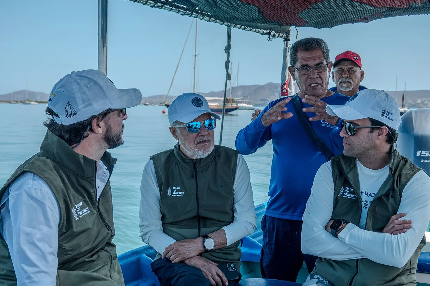 Group of five men sitting on a boat, engaging in conversation, with a calm bay and sailboats in the background.