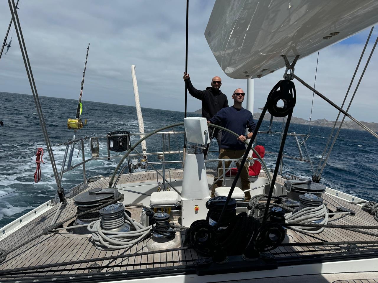 Two men standing on a sailboat at sea, one holding a rope and the other looking forward, with a scenic ocean view in the background.