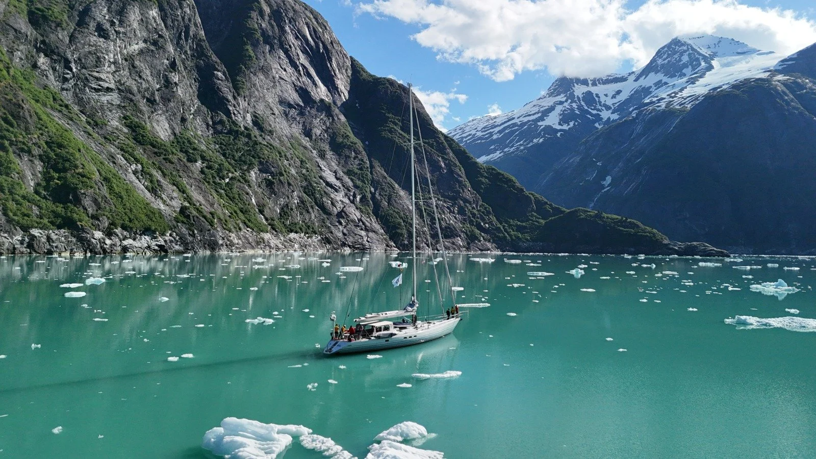 A sailboat floating on icy turquoise water with floating ice chunks, surrounded by steep green mountains and snow-capped peaks under a partly cloudy sky.
