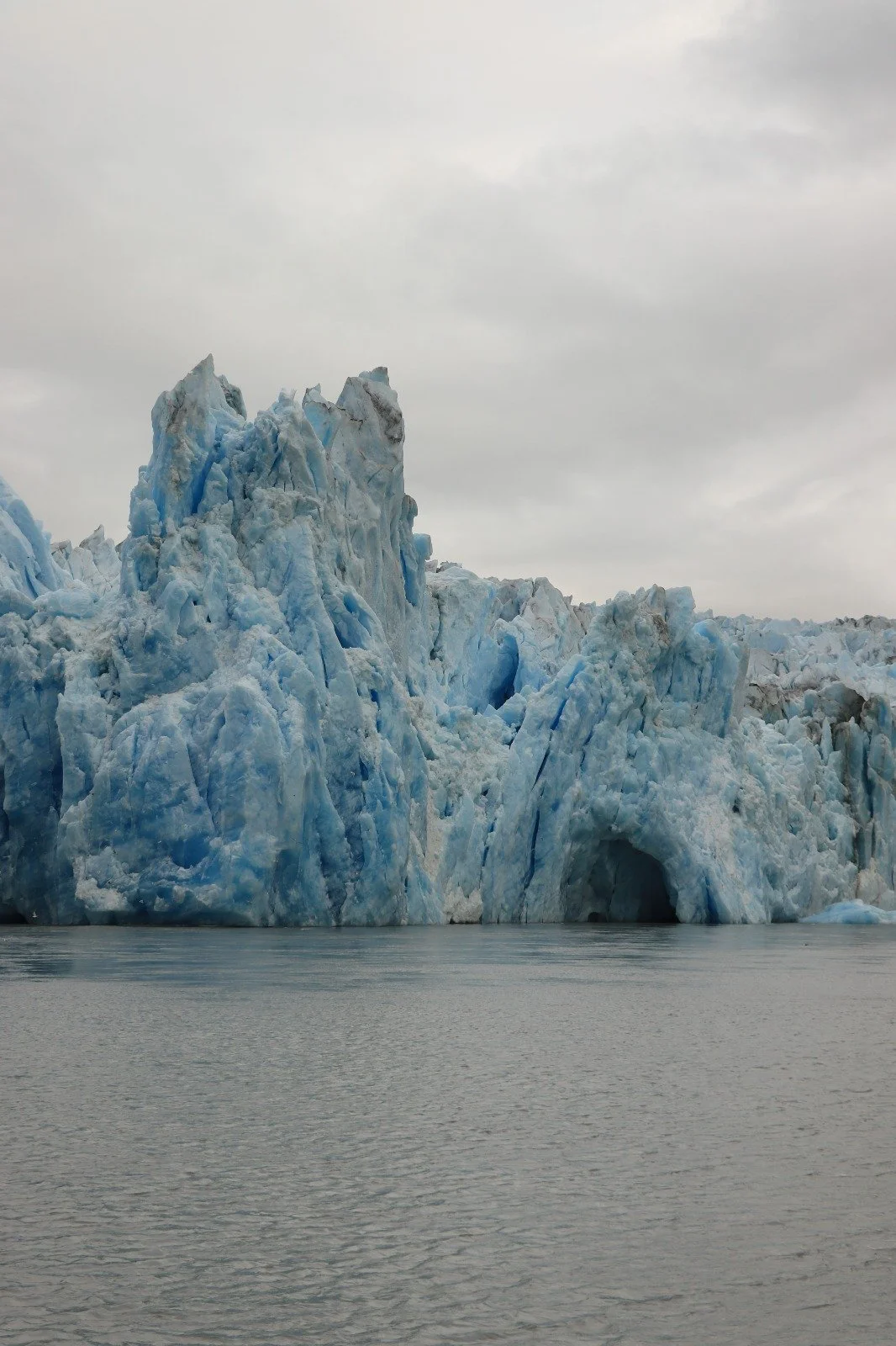 A large glacier with jagged ice formations extending into a calm body of water, under a cloudy sky.