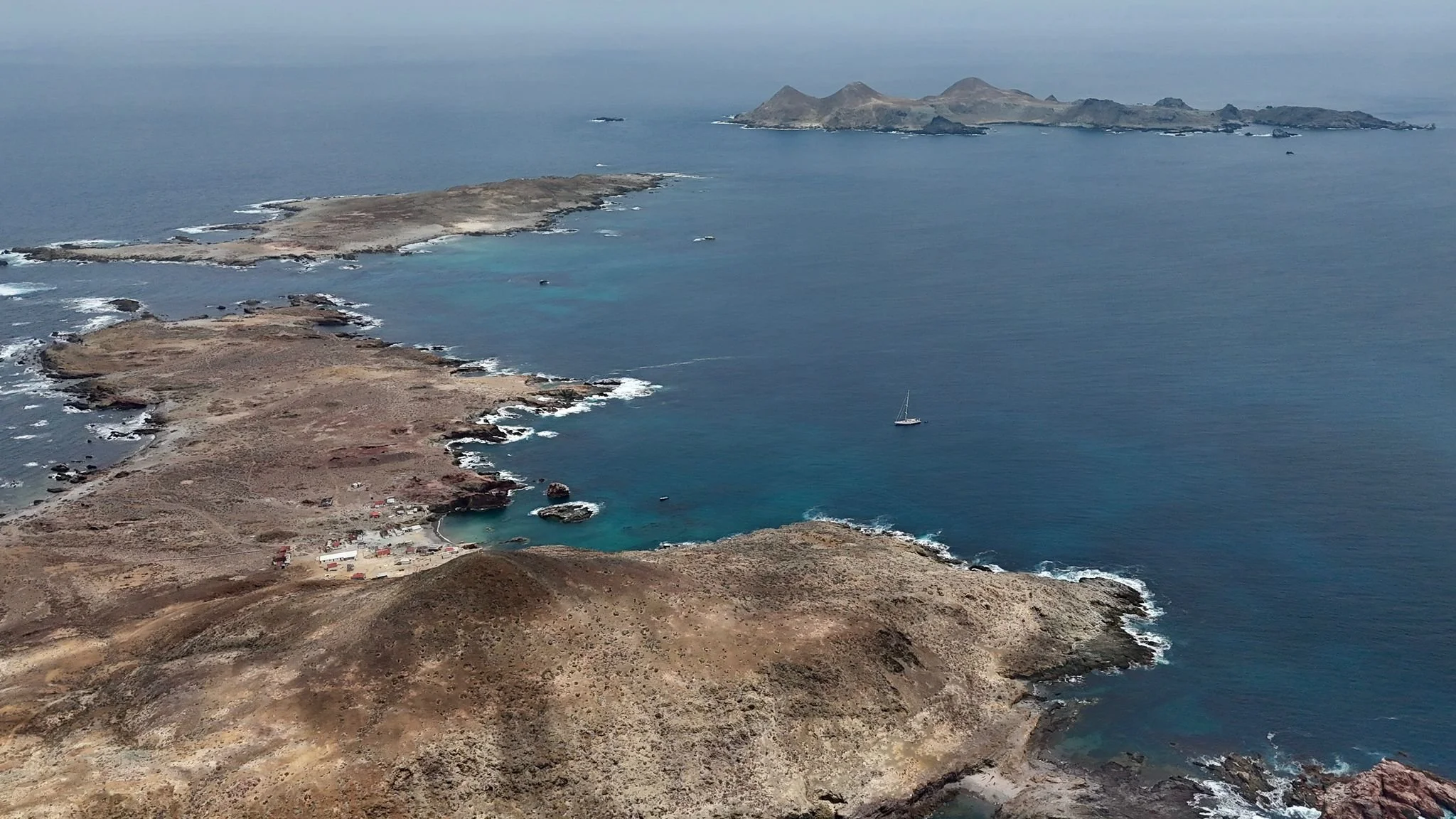 Aerial view of a rugged coastal landscape with a small harbor, rocky shoreline, and distant islands out at sea.