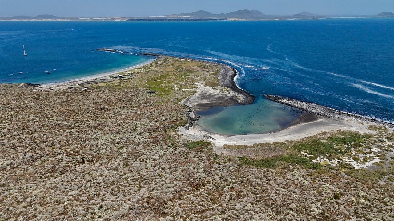 Aerial view of a coastal area with a small lagoon, a sandy beach, and a curved seawall protecting the shoreline, with boats sailing in the ocean and distant mountains on the horizon.