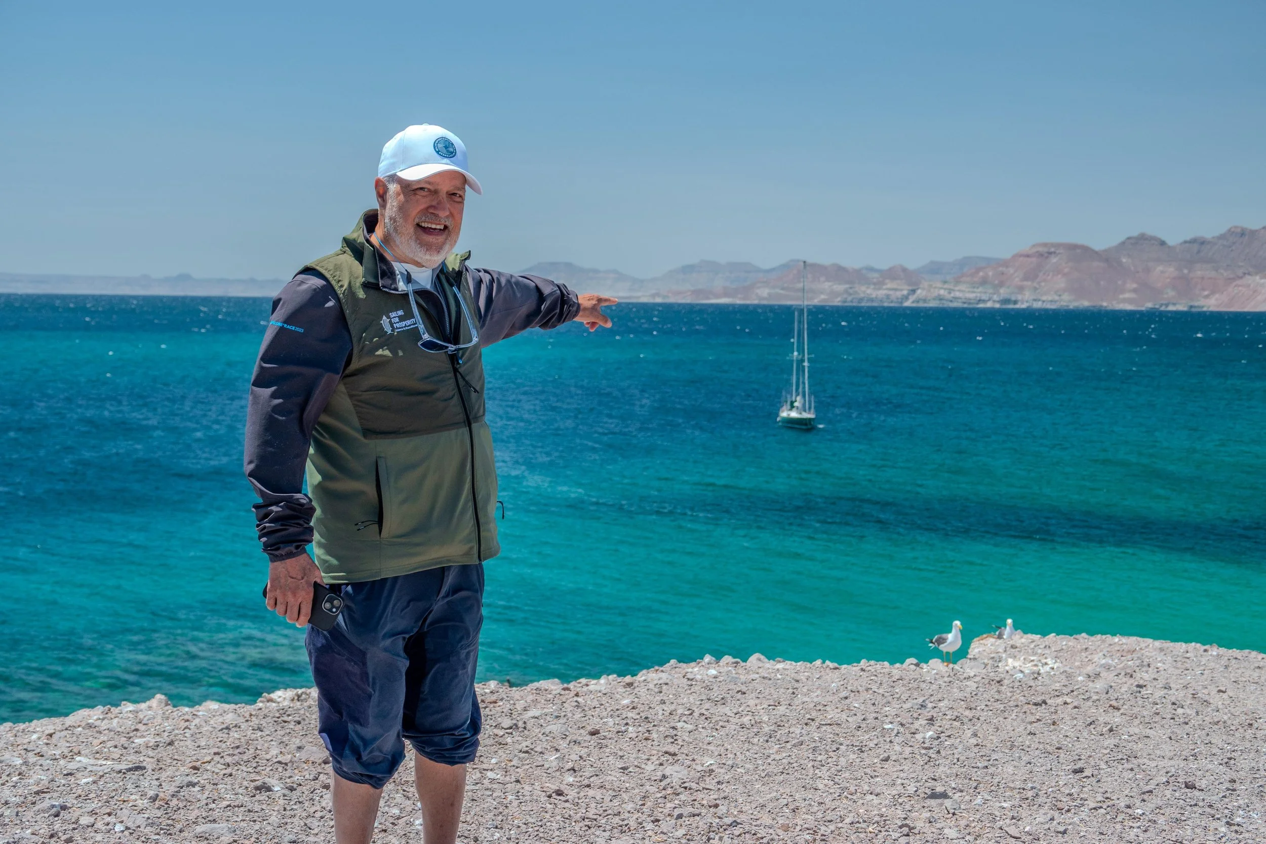 A man smiling and pointing towards the ocean on a sunny day with a sailboat in the background, seagulls on the rocky shore nearby.