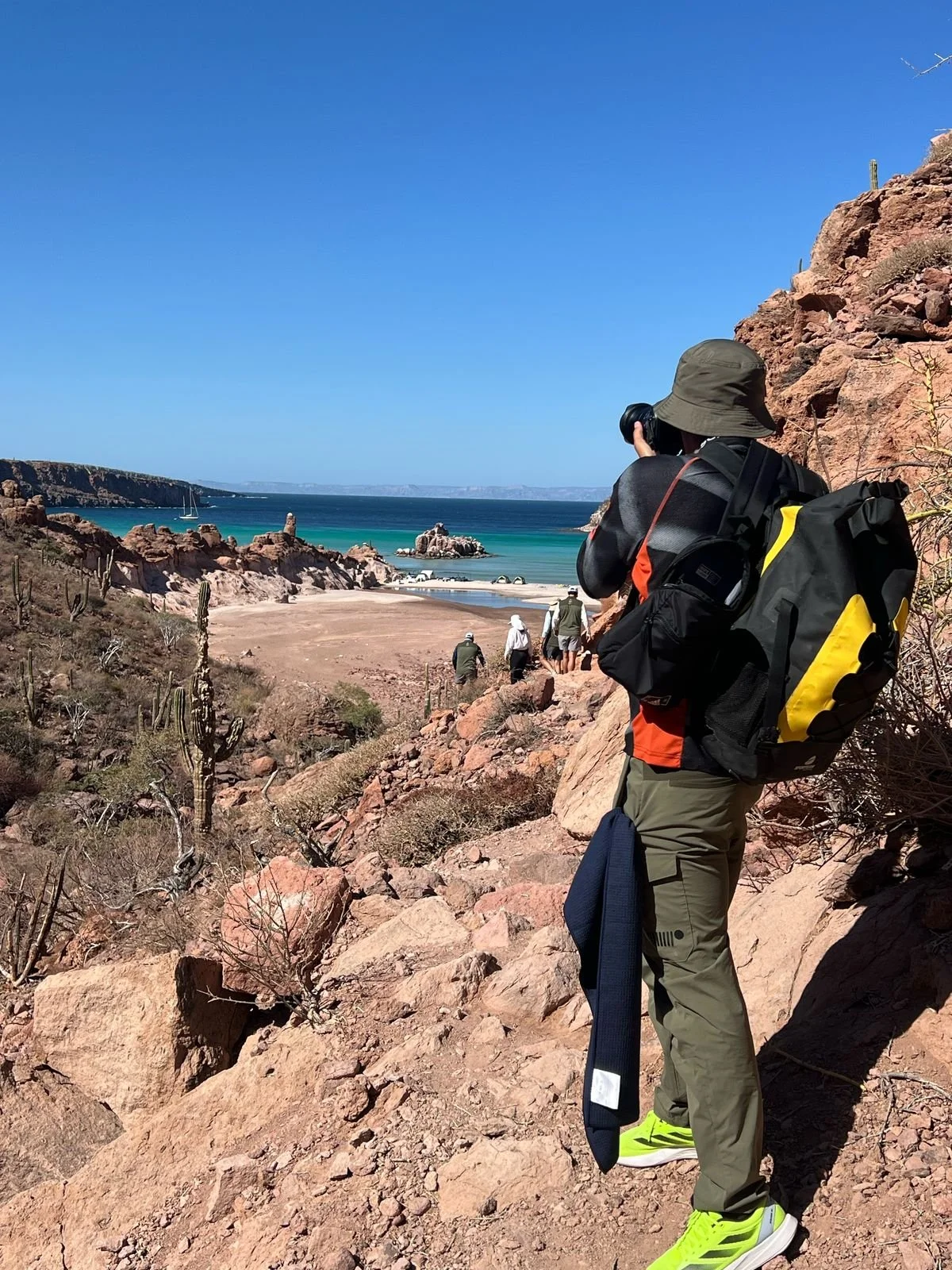 Person hiking on rocky terrain with a backpack and camera, overlooking a sandy beach, rocky formations, and cacti with a clear blue sky.