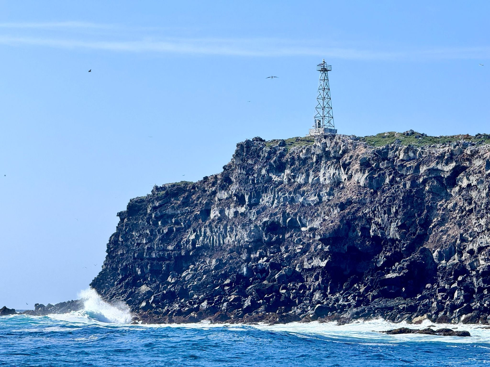A rocky cliff with a lighthouse on top, ocean waves crashing against the rocks, and a clear blue sky with some birds flying.