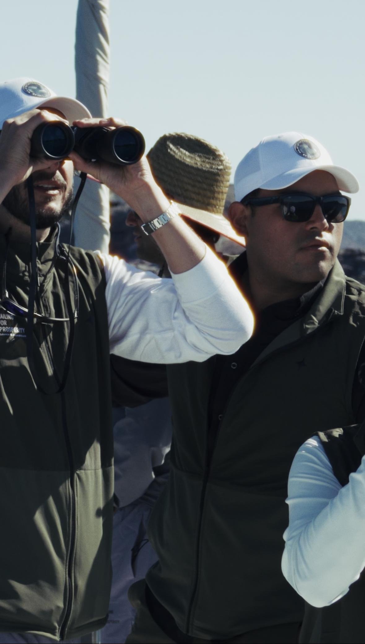 Two men wearing sunglasses and hats, one looking through binoculars, outdoors on a sunny day.
